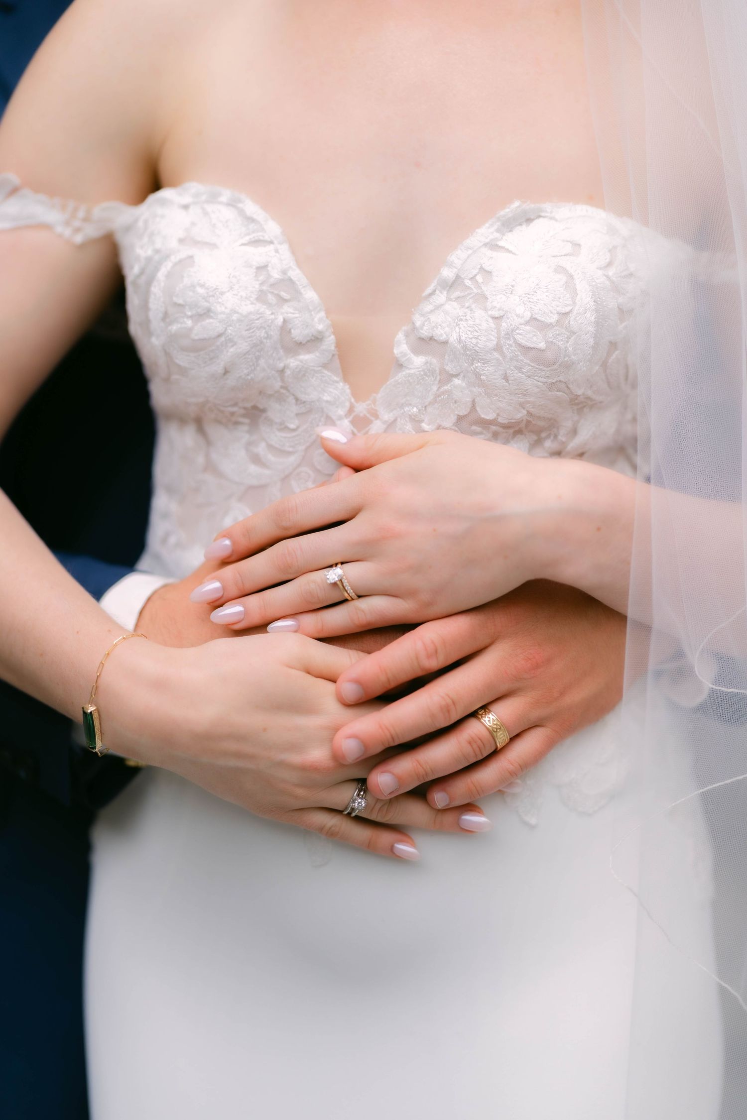 Bride and groom hands with wedding rings clasped together over white lace bridal gown.