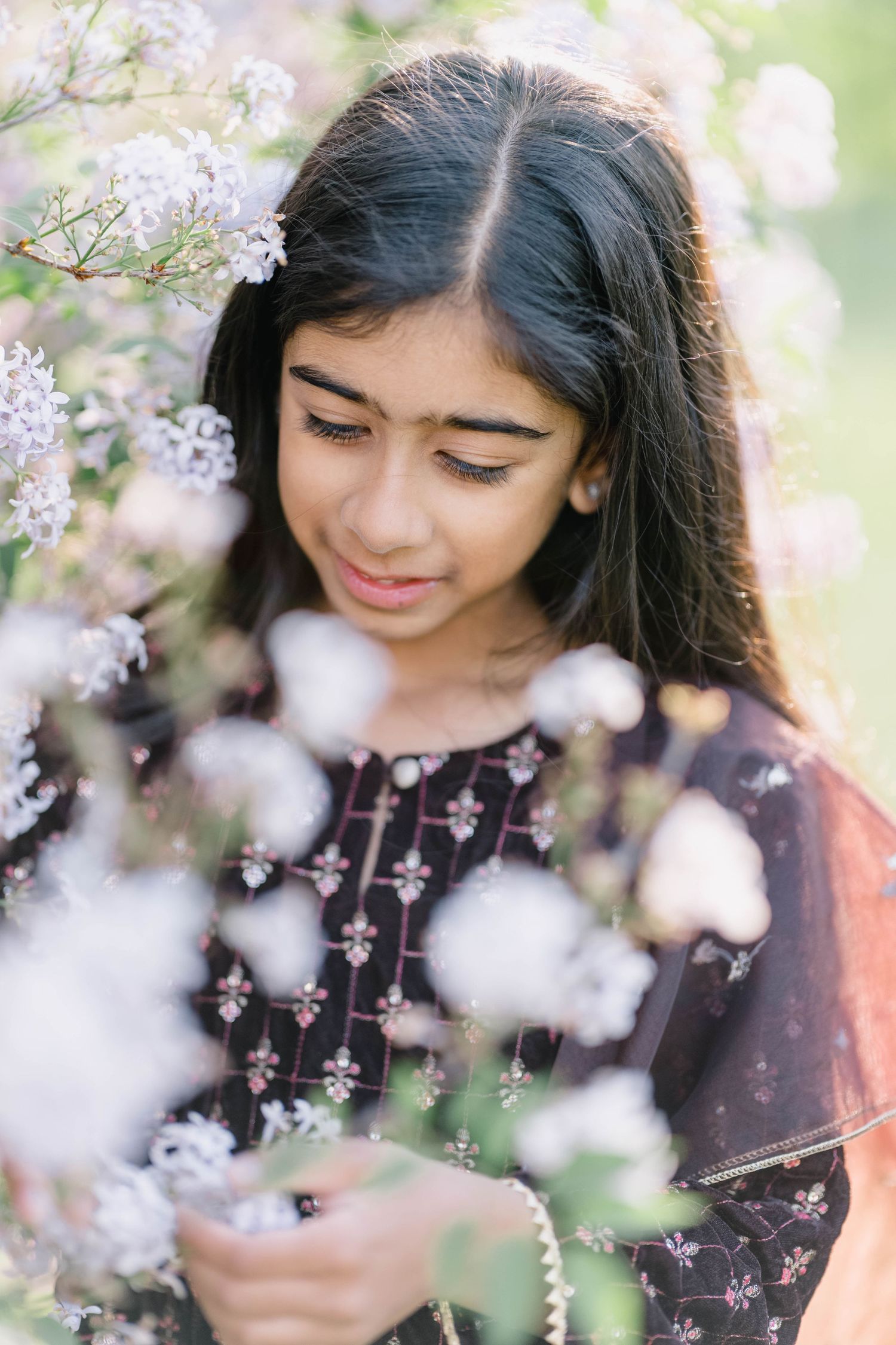 Young South Asian girl in embroidered dark dress surrounded by white blooming flowers, looking down peacefully.