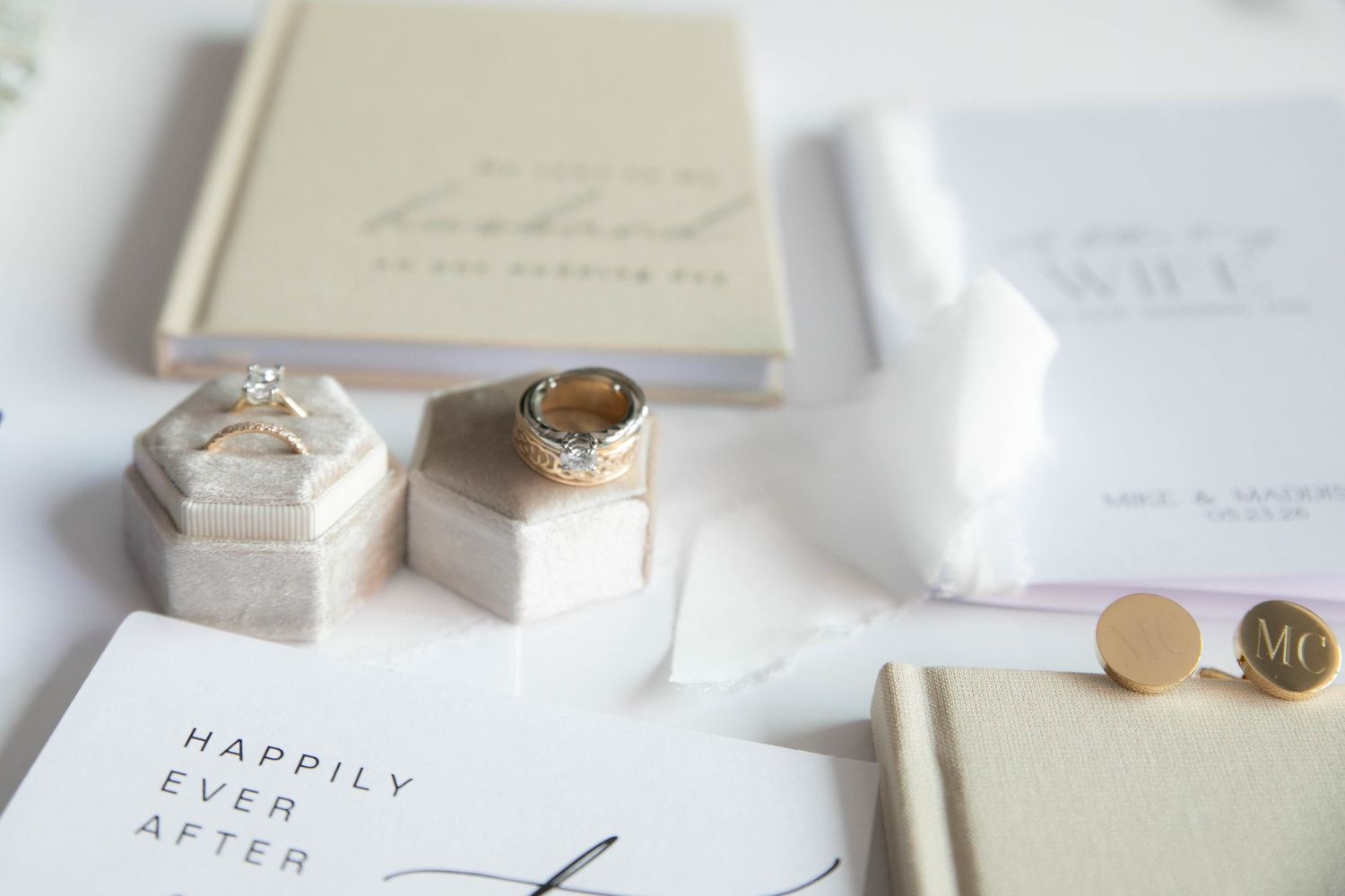 Elegant wedding flat lay with diamond engagement ring, gold band, cufflinks, and stationery on white background.