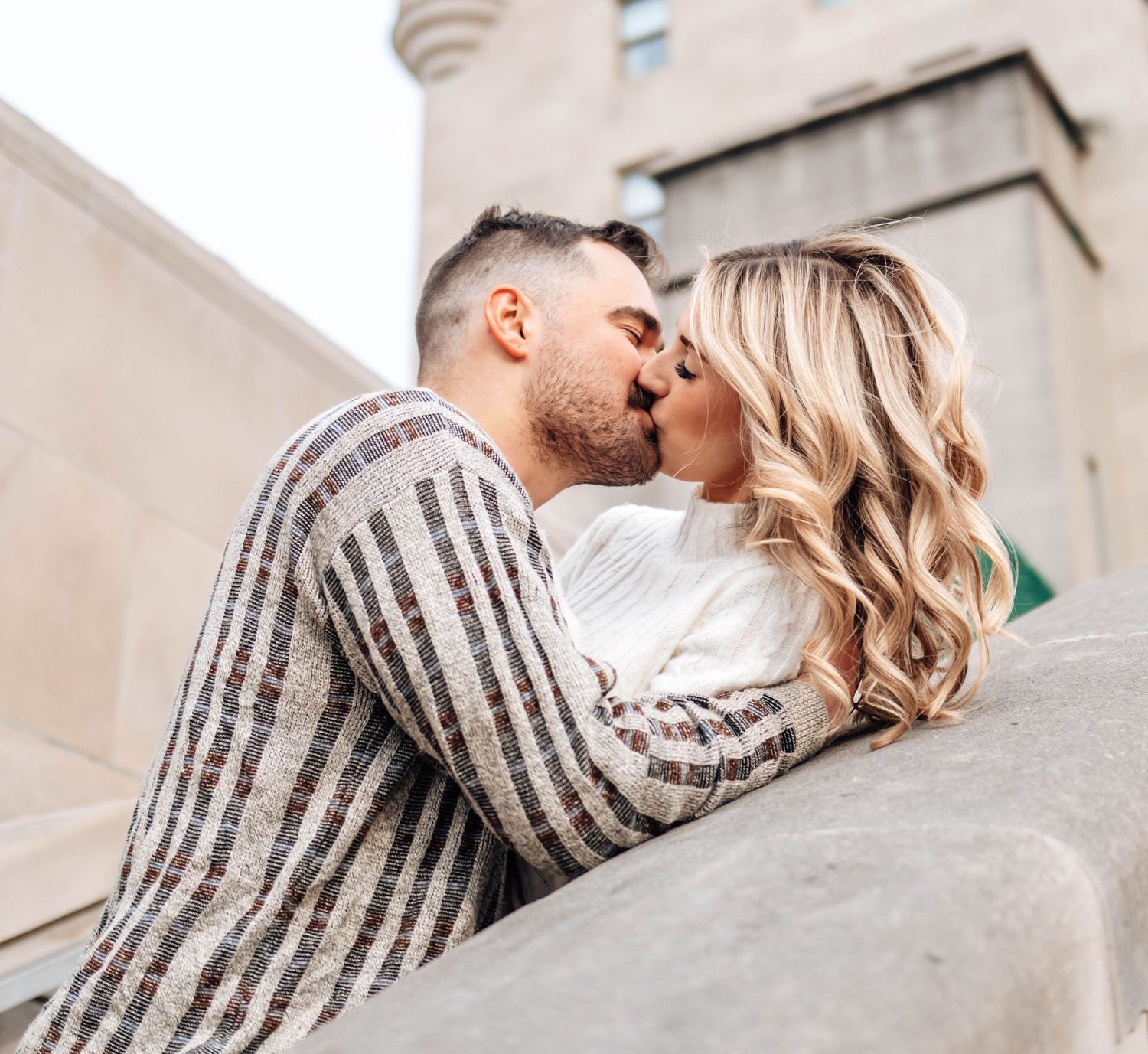 A couple in a striped sweater and white dress share a romantic kiss against a beige wall backdrop.