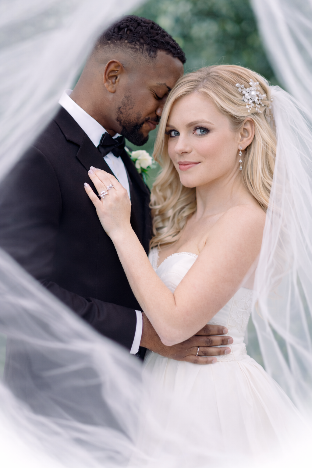 A romantic wedding portrait of a couple embracing under a flowing veil in an outdoor garden setting.