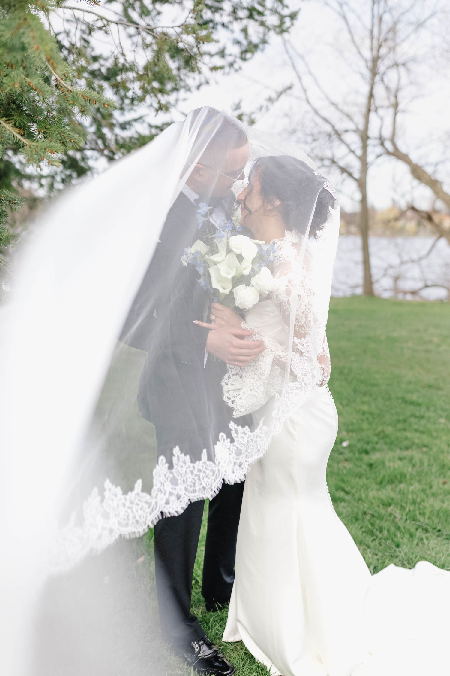 Bride and groom kissing under flowing lace-trimmed veil outdoors near lake holding white bouquet.