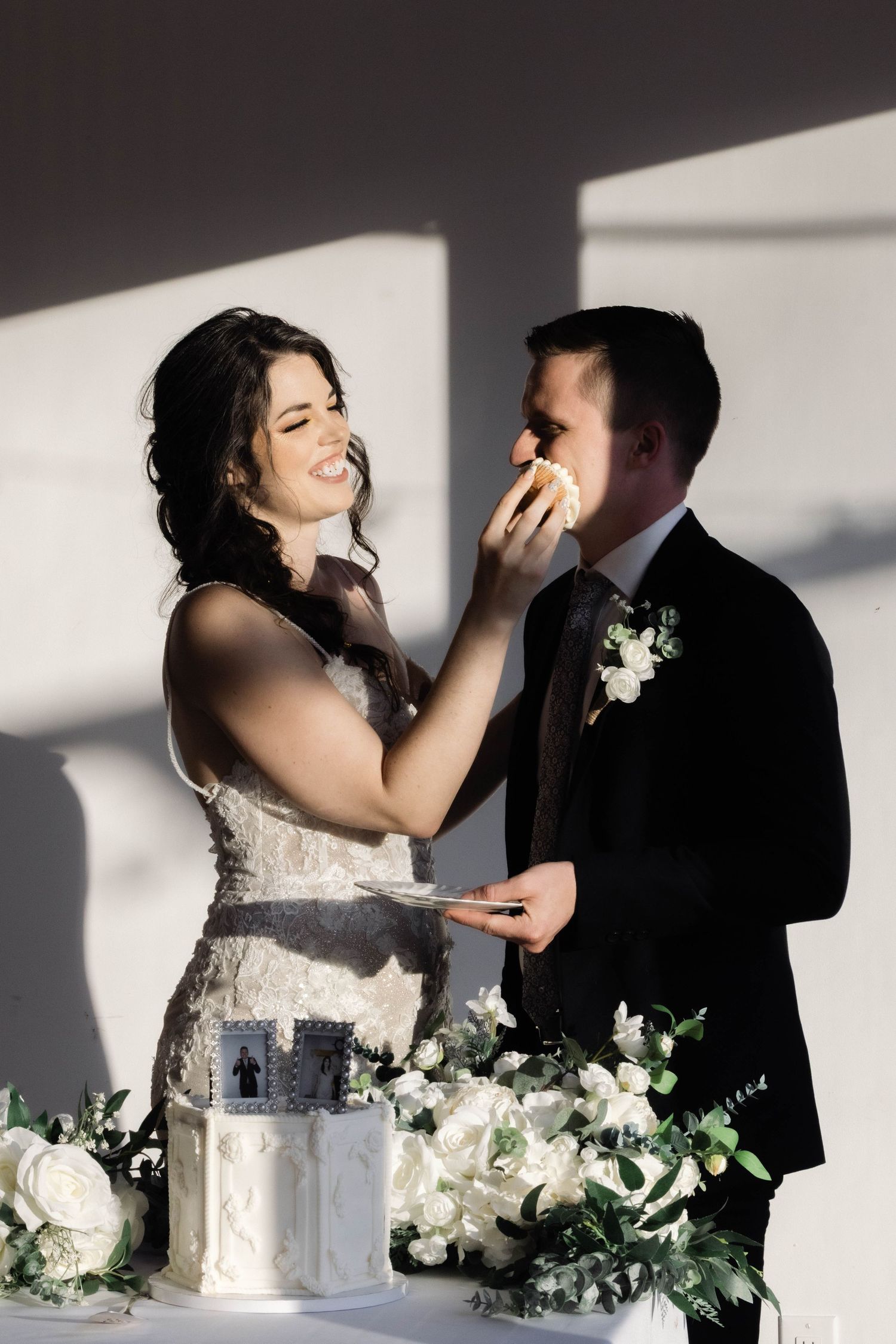 Newlyweds share a playful moment while cutting their elegant white wedding cake adorned with flowers.
