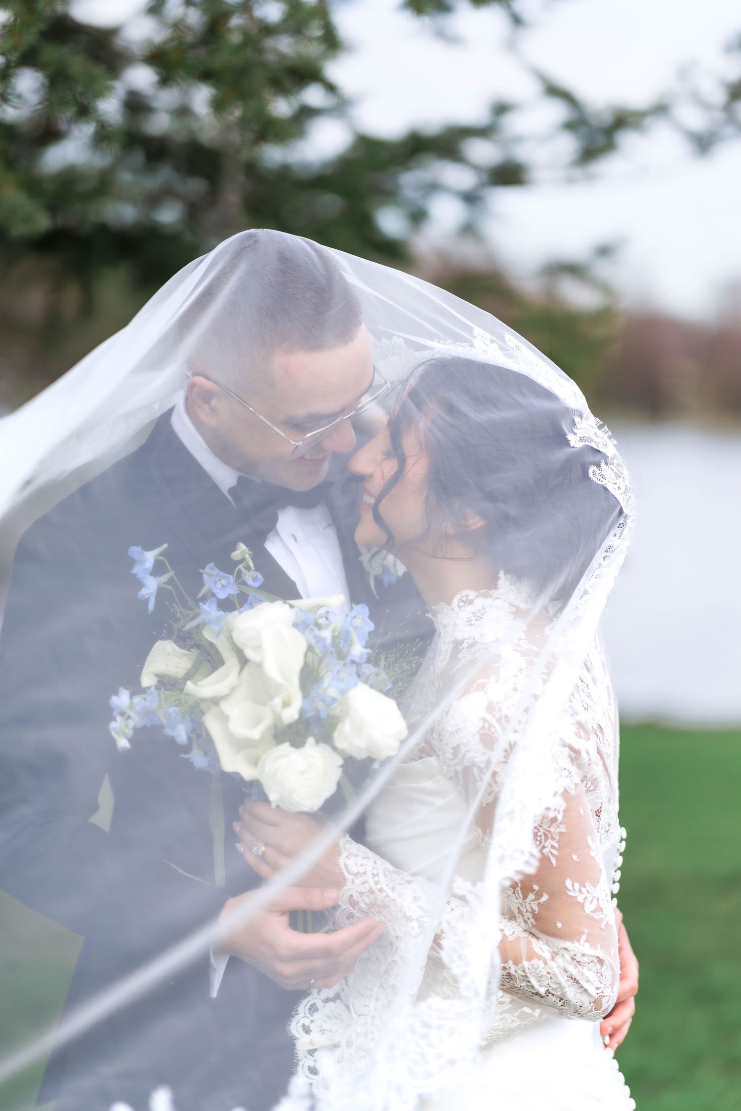 Bride and groom share a romantic kiss under flowing white veil, holding white and blue floral bouquet outdoors.