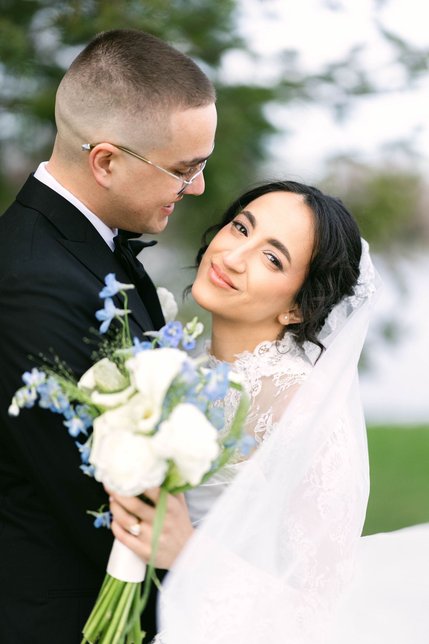Bride and groom embrace outdoors, bride smiling in lace veil holding white and blue floral bouquet.