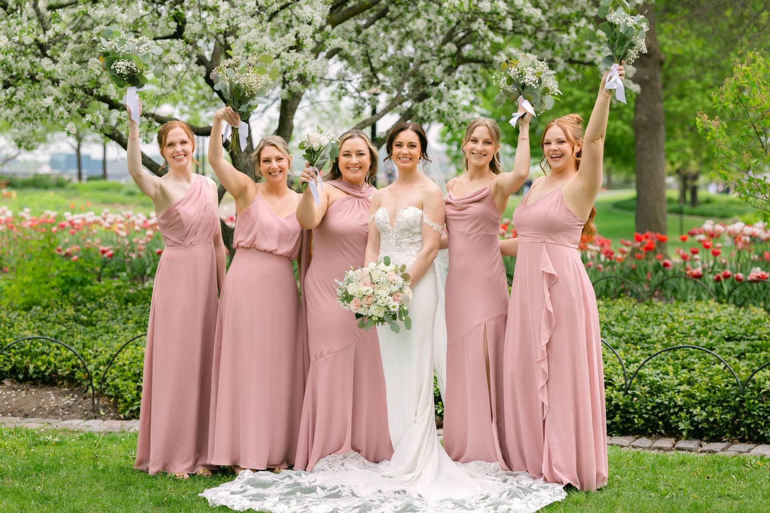 Bride and four bridesmaids in dusty rose gowns celebrate outdoors among blooming trees and red tulips.