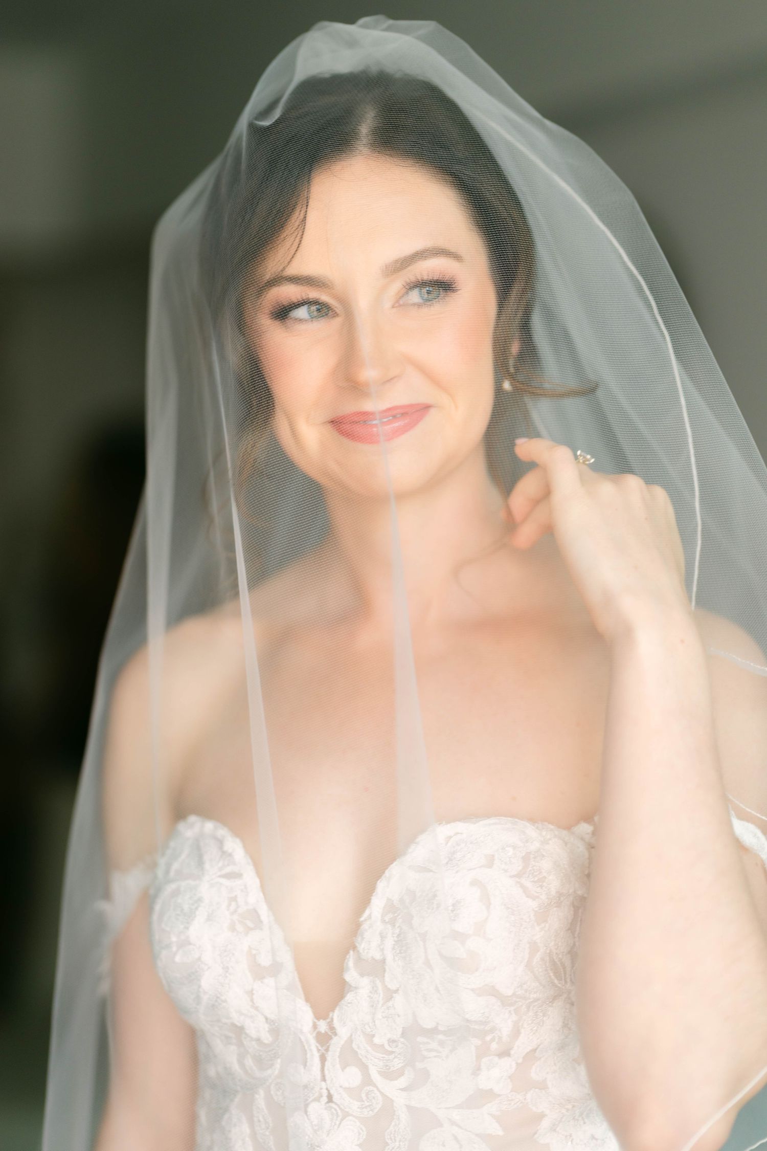 Bride in lace strapless gown and sheer veil smiles softly, touching her earring in an elegant bridal portrait.