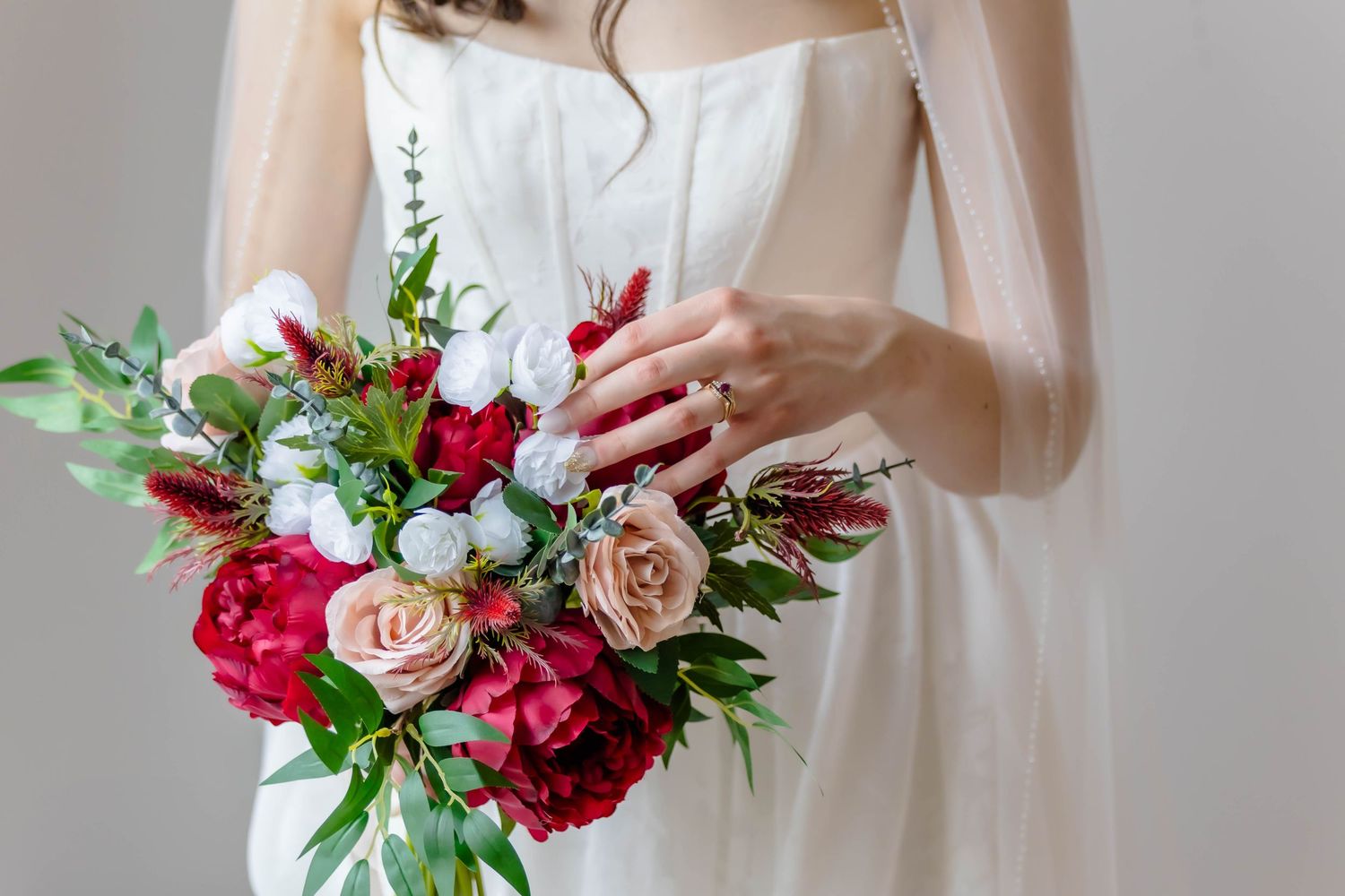Elegant bridal bouquet featuring red and pink peonies, blush roses, and greenery held against a white wedding dress.