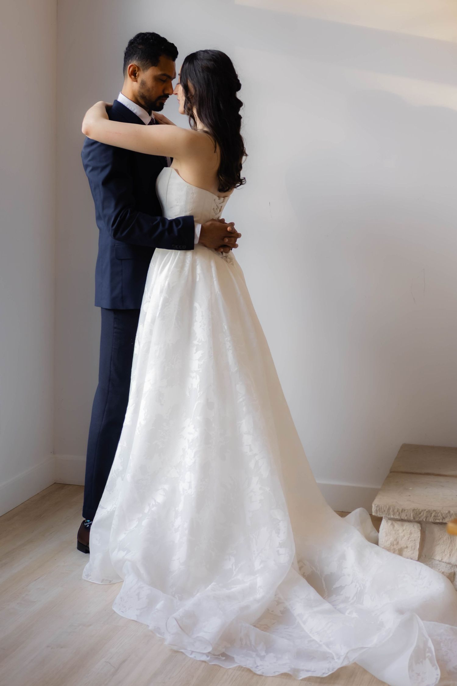 Elegant couple in navy suit and white lace wedding gown dancing in a bright indoor setting.