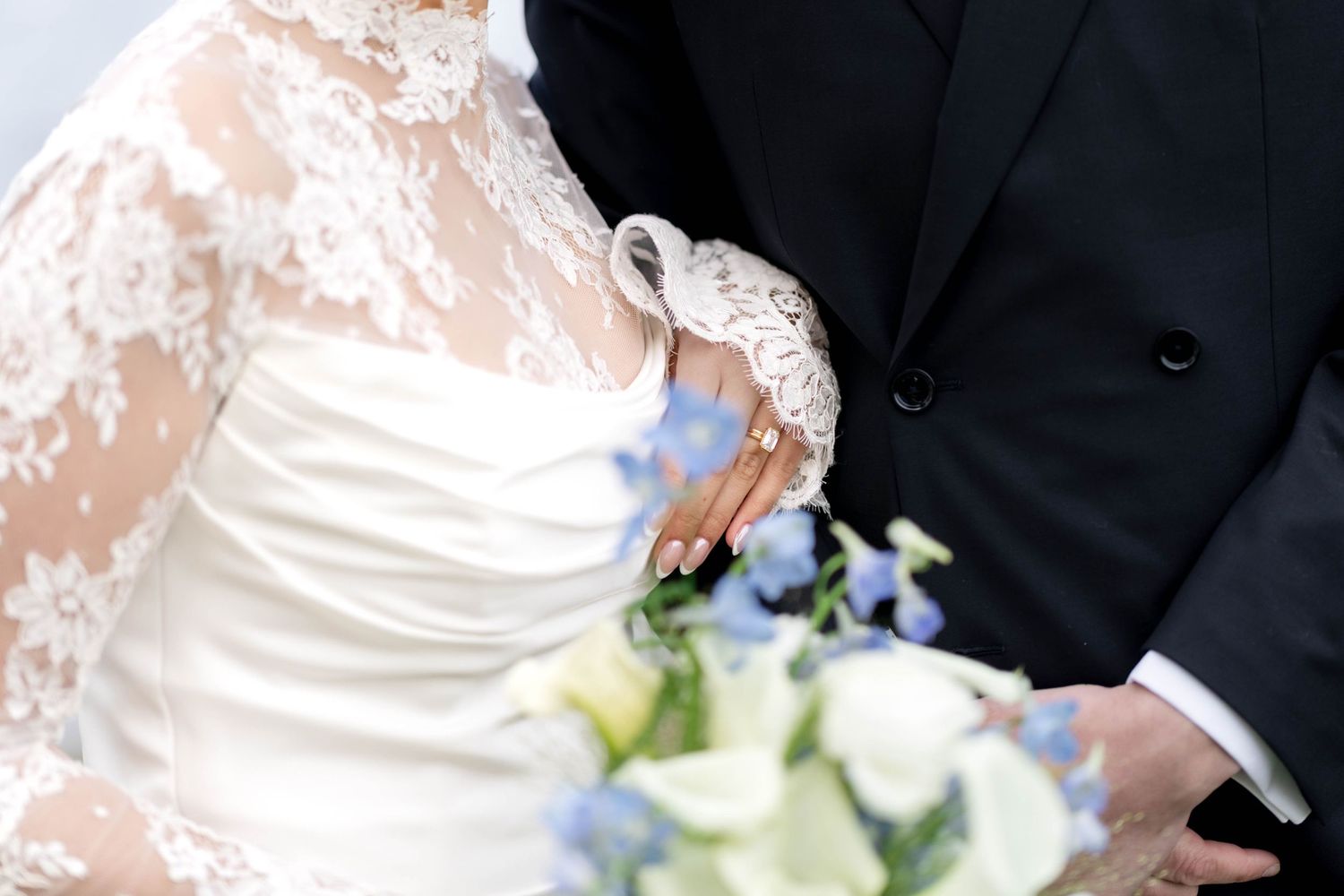 Bride in lace wedding dress holding blue and white bouquet beside groom in black suit at wedding ceremony.
