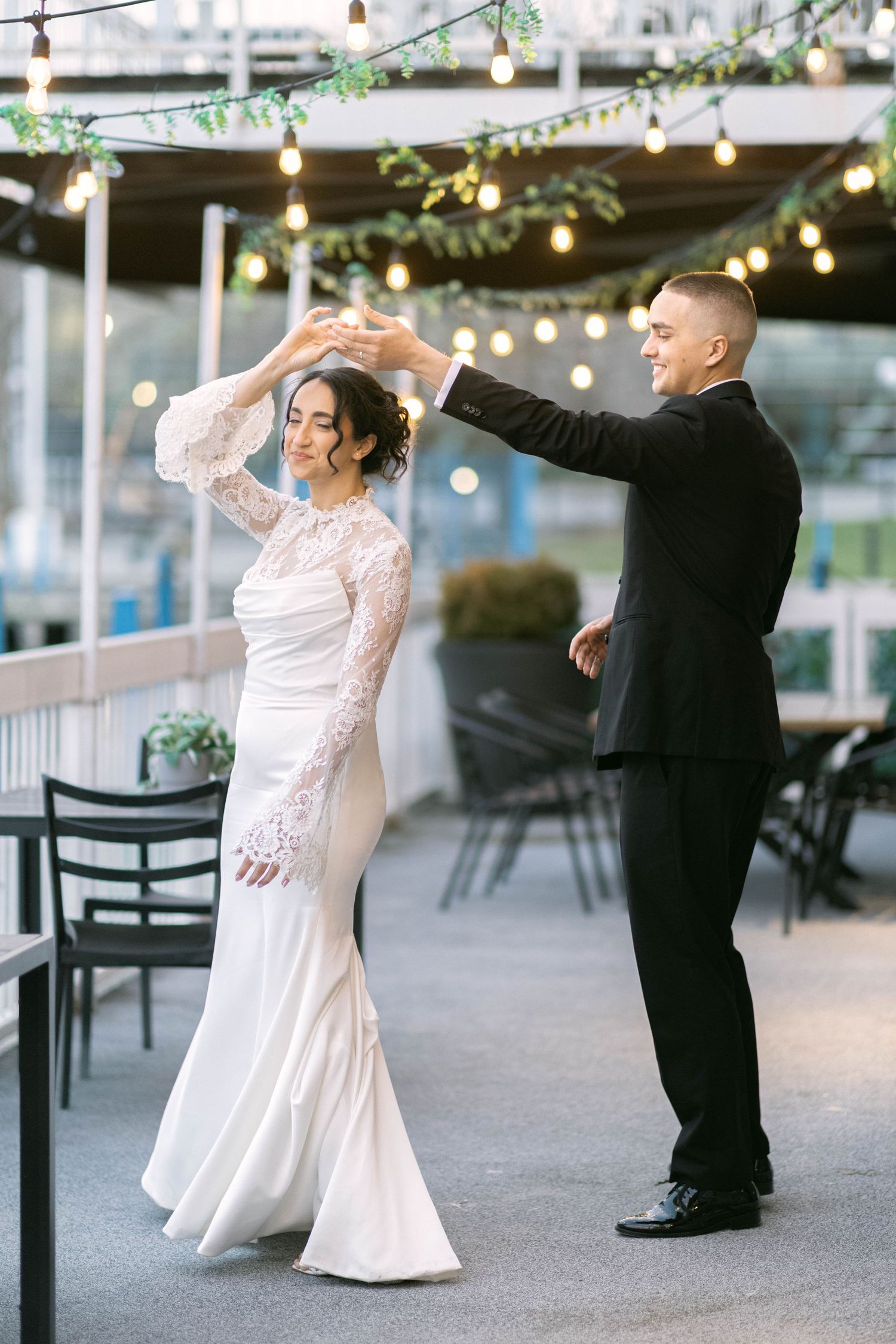 Bride in lace gown twirls as groom in black suit leads first dance on outdoor terrace with string lights.