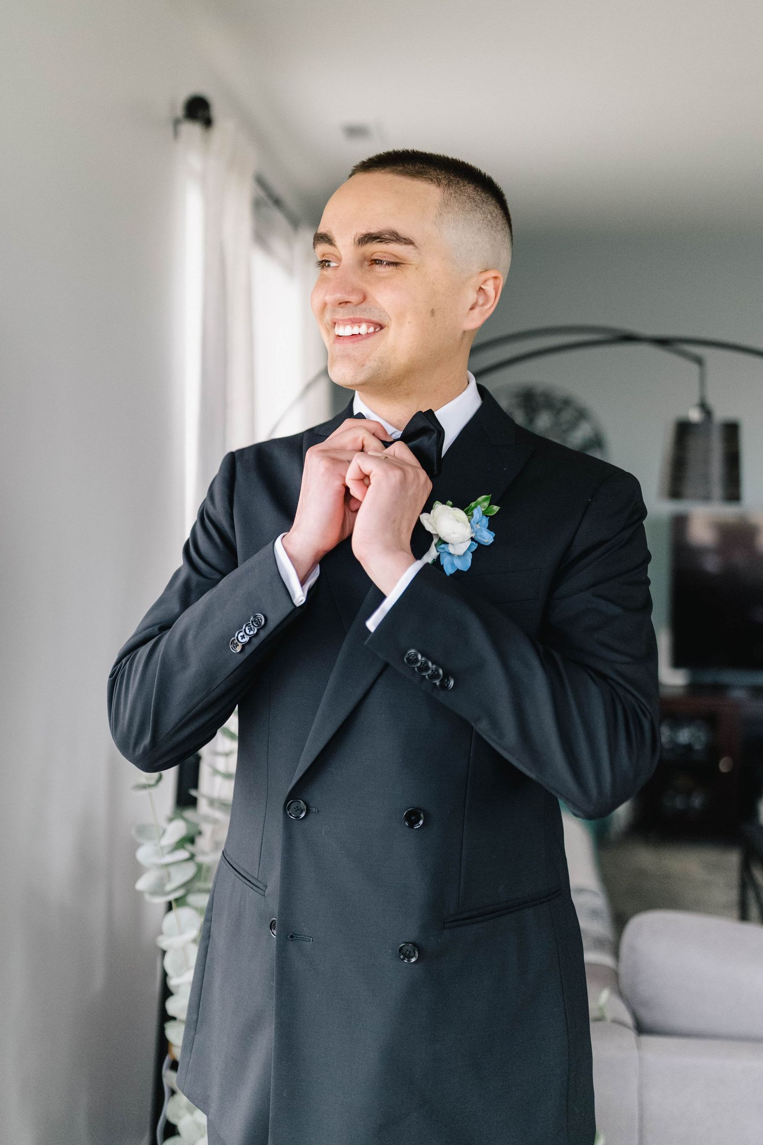Smiling groom in black double-breasted tuxedo adjusting bow tie, wearing white floral boutonniere, standing indoors.