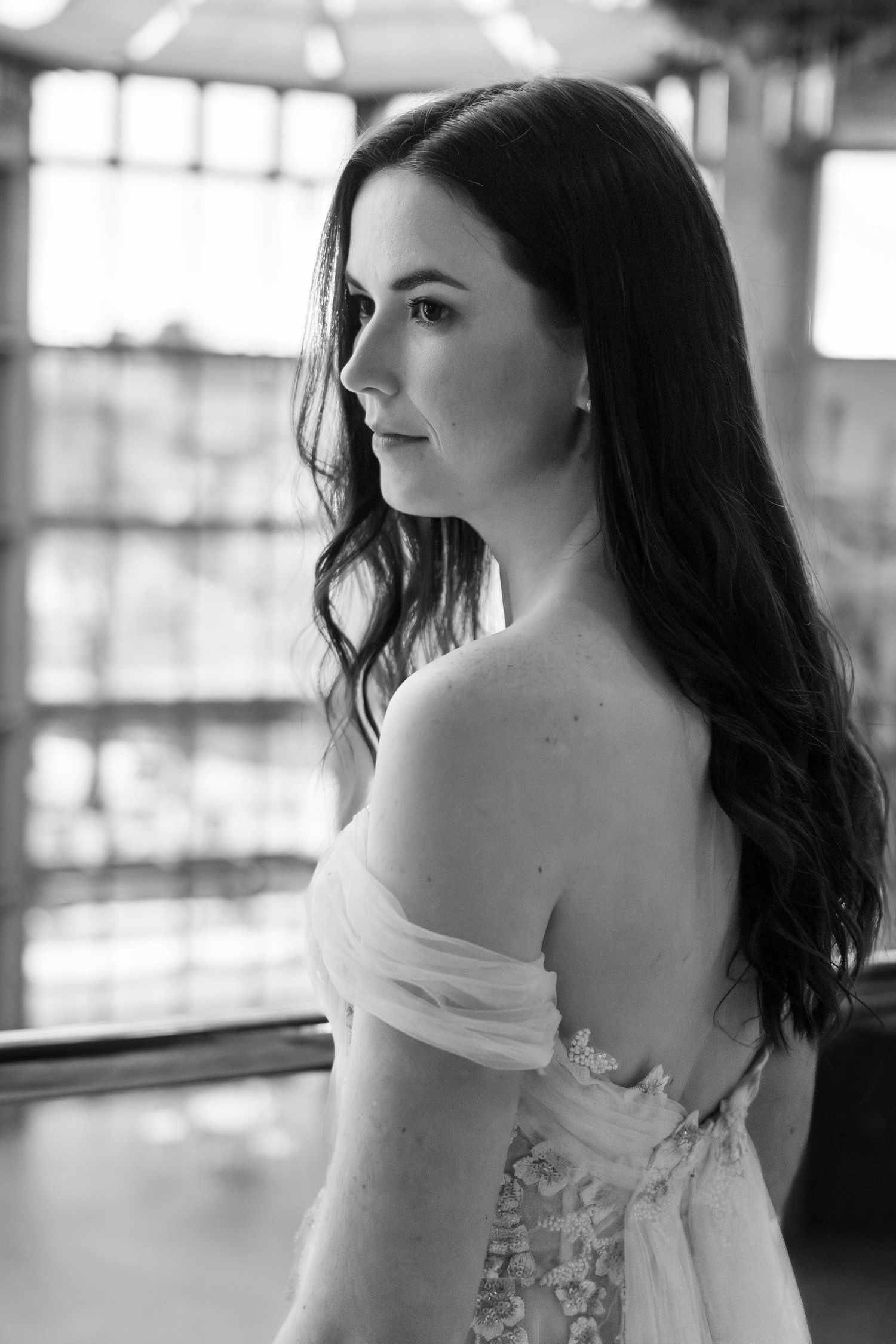 A black and white portrait of a woman in an off-shoulder lace wedding dress looking to the side in soft natural light.