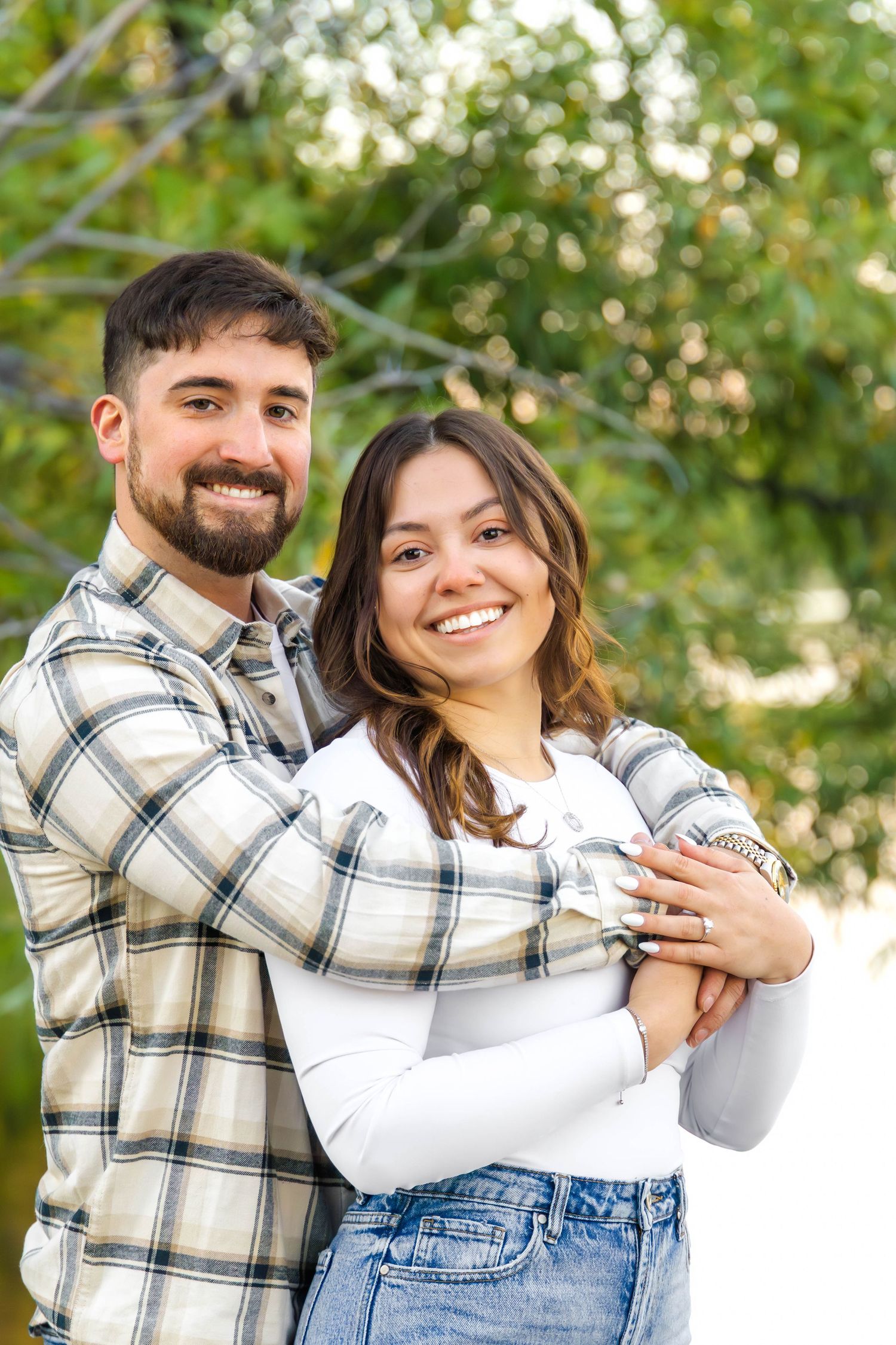 Playful couple embracing and laughing together in front of lush green trees during outdoor photo session.
