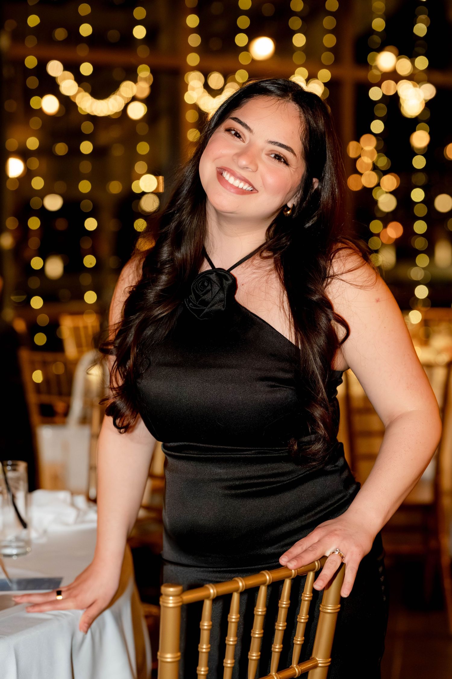 A smiling woman in a black halter dress poses by a gold chair at an elegant event with bokeh lights.