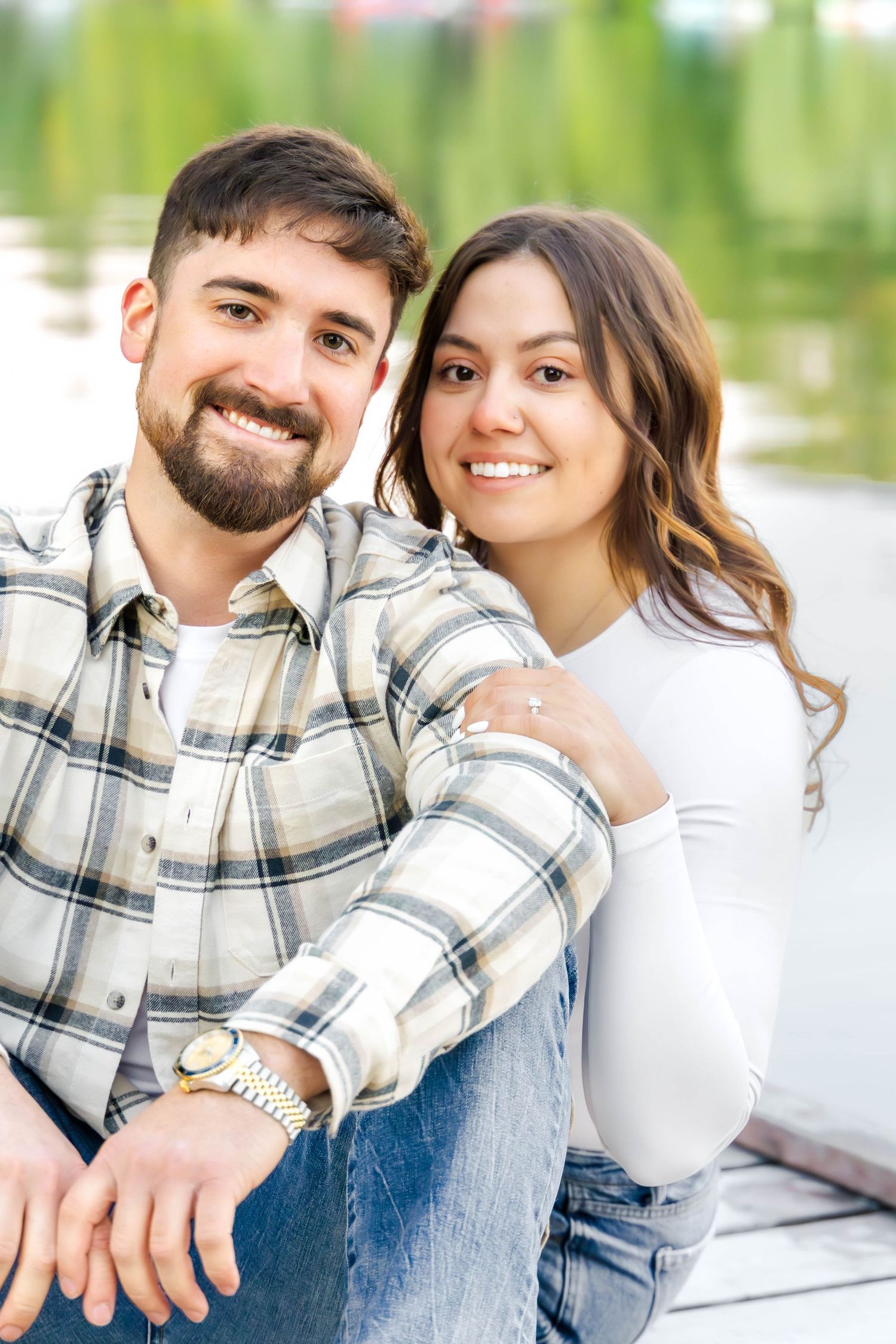 Close-up of smiling couple in casual wear against blurred lakeside setting.