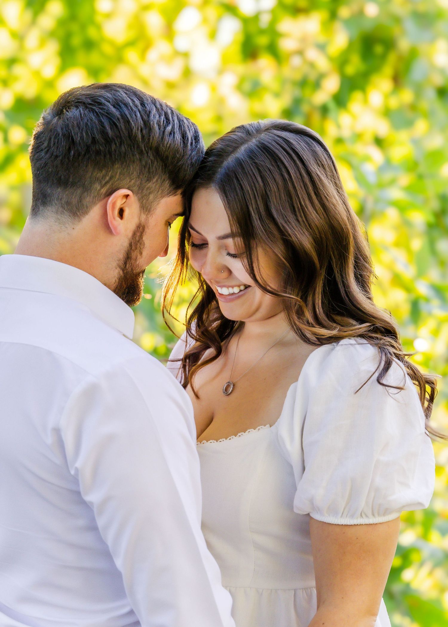 Tender moment between couple in white outfits against bright green foliage background.