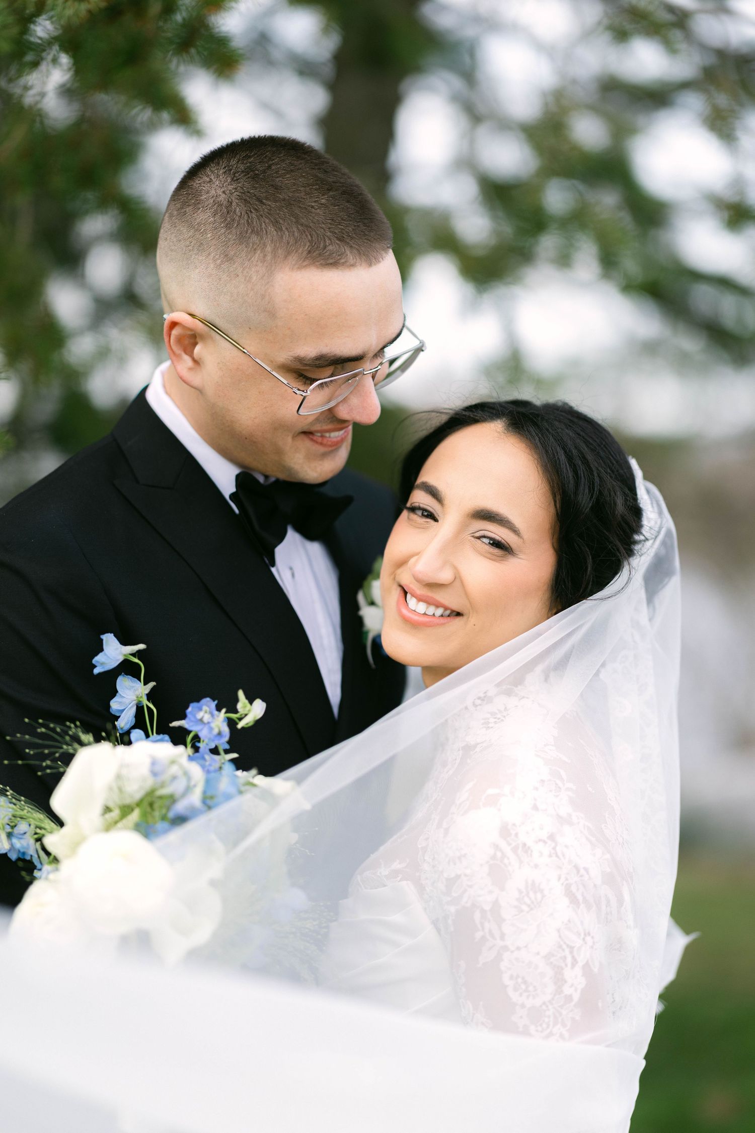 Smiling bride in lace gown and veil holds blue and white bouquet while groom in black tuxedo gazes at her outdoors.