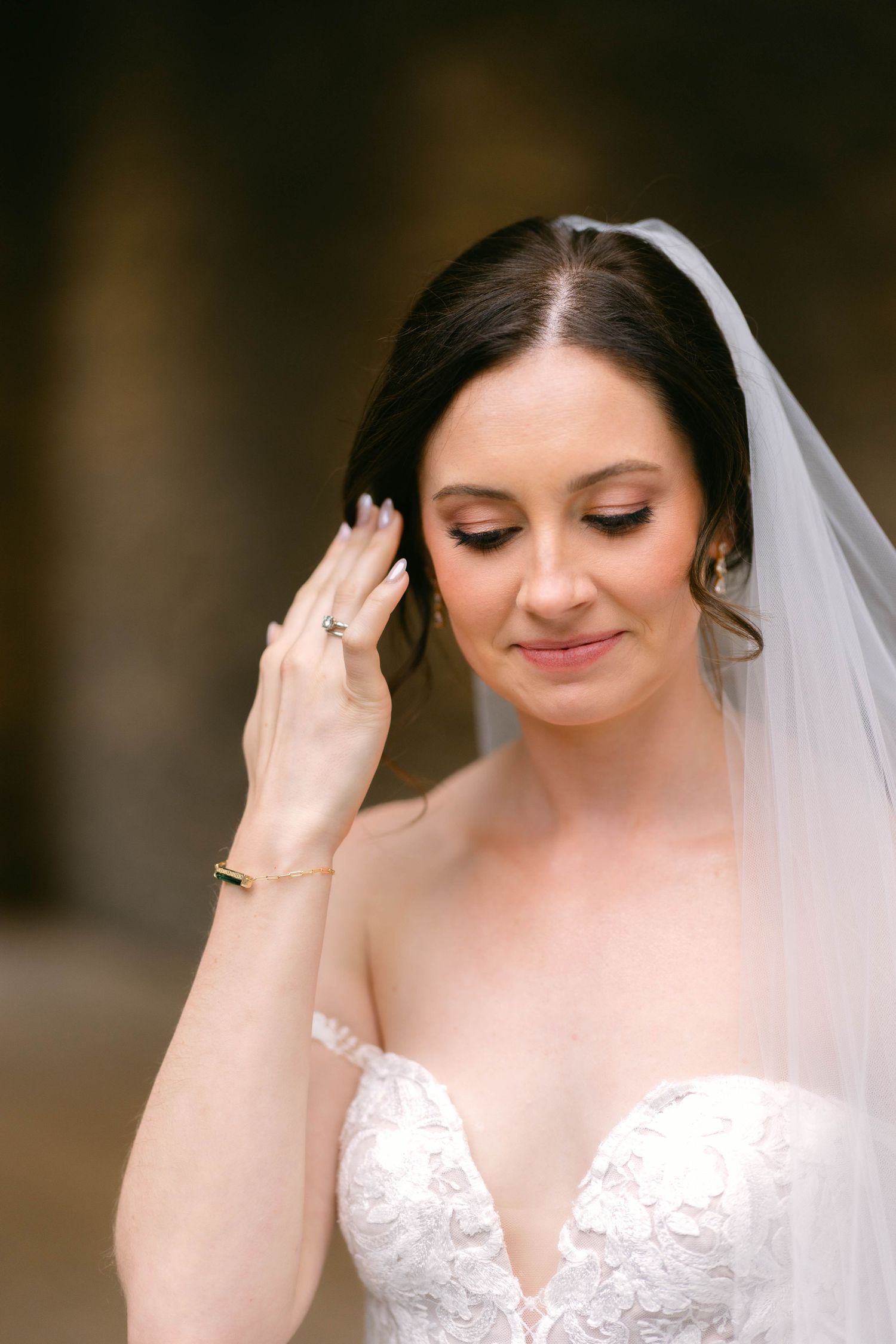 Beautiful bride in lace wedding gown and veil looks down gracefully, showcasing her engagement ring and bracelet.