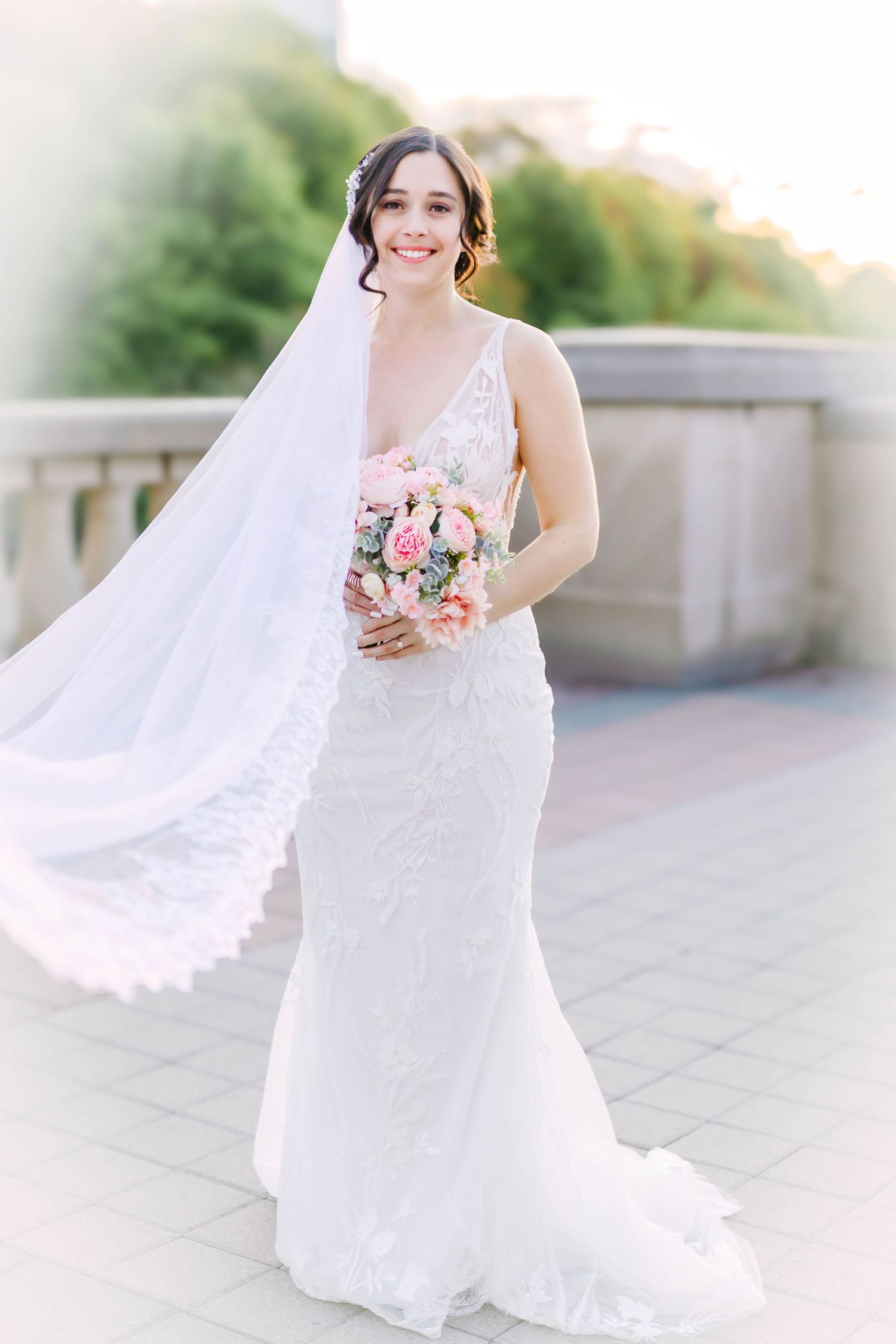 A bride in a fitted lace wedding dress poses outdoors with a cathedral veil and pink bouquet.