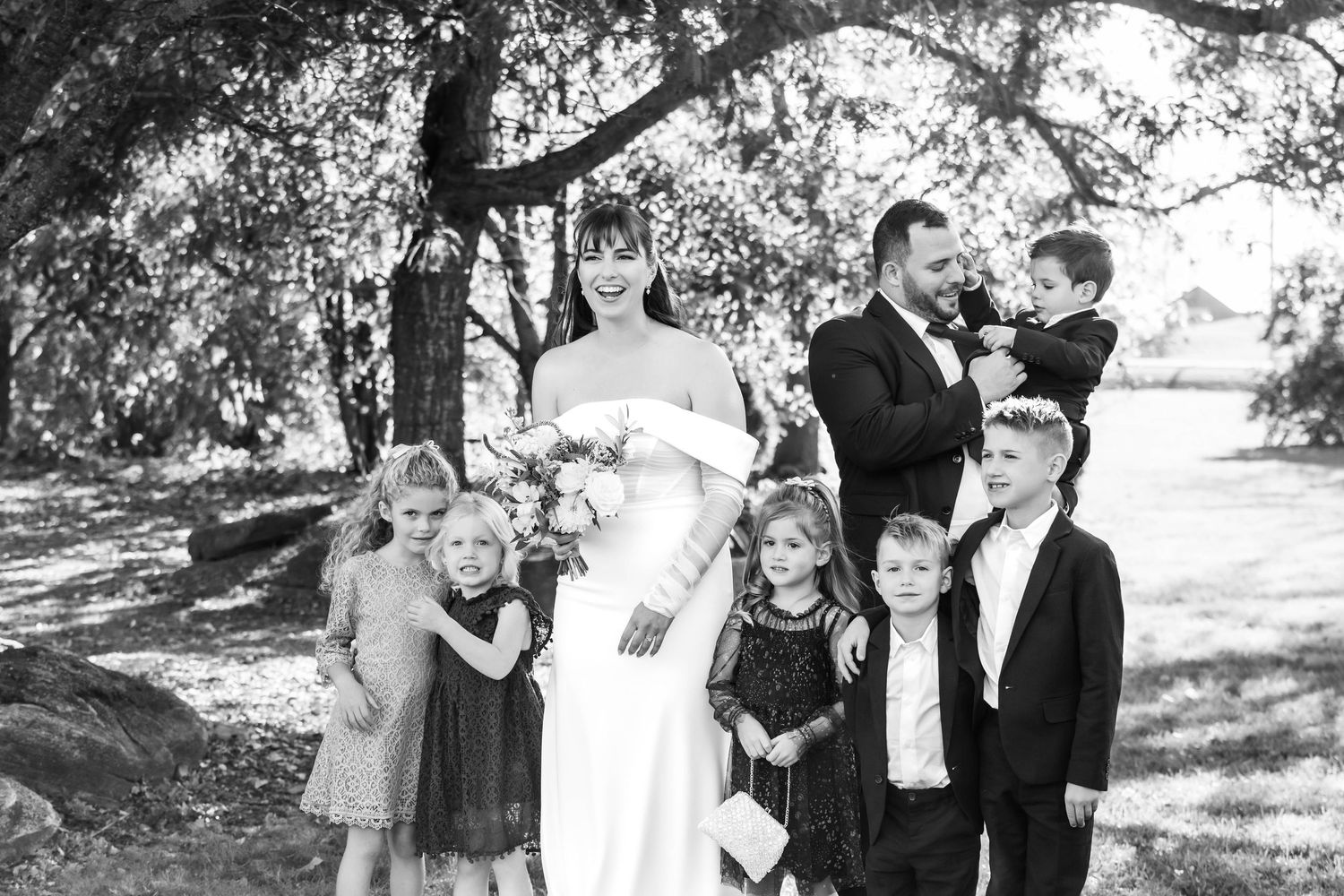 Wedding party poses for formal portraits under shaded trees outdoors.