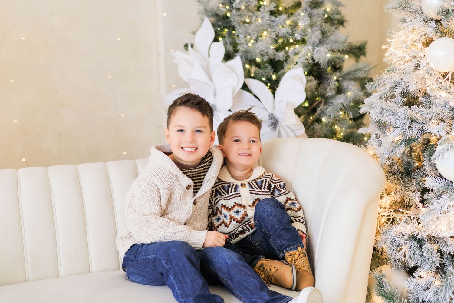 Siblings sitting together on white couch with decorated Christmas tree in background.