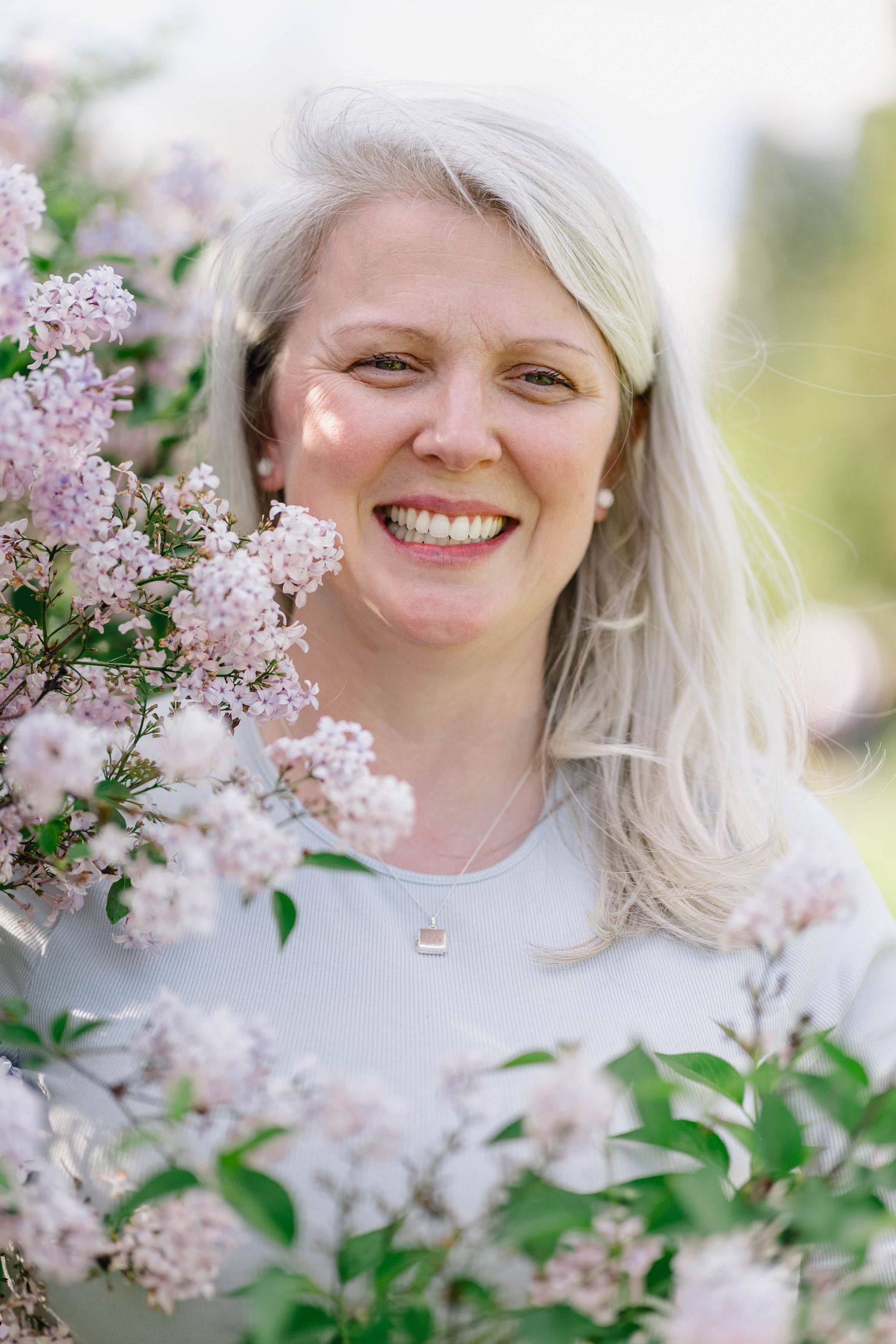 Smiling middle-aged woman with silver hair surrounded by blooming pink lilac flowers outdoors.