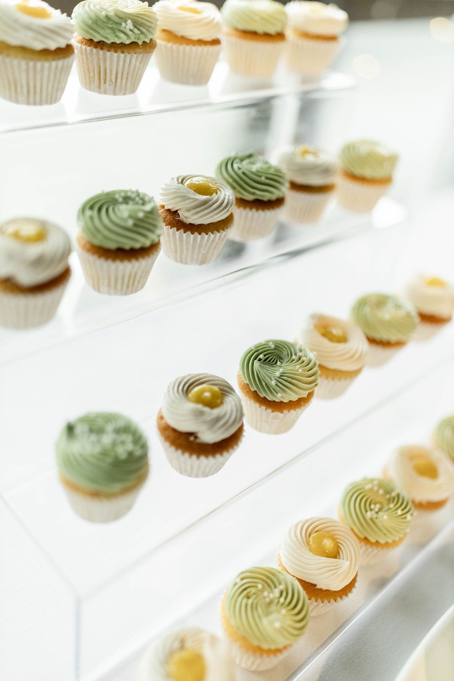 Elegant display of green and white frosted cupcakes with gold pearl decorations arranged on clear acrylic shelves.