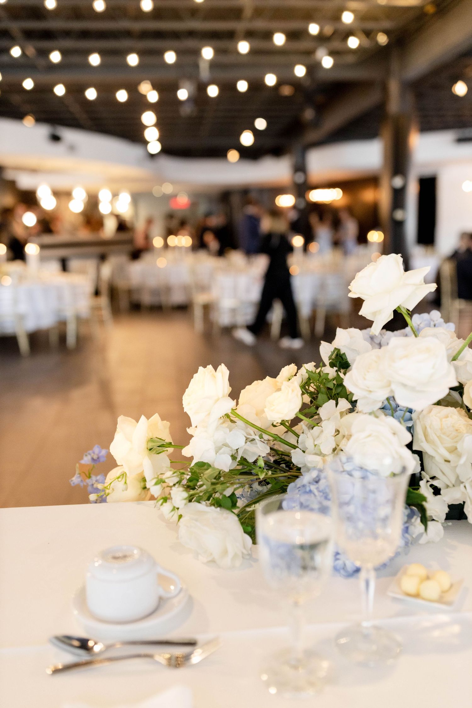 Bright wedding reception hall with string lights, white and blue floral centerpieces on draped tables.
