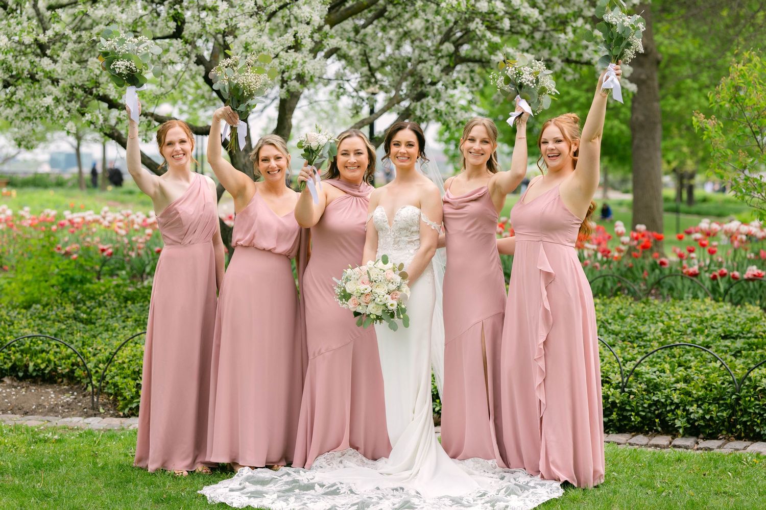 Bride and four bridesmaids in dusty rose gowns celebrate outdoors among blooming trees and colorful tulips.