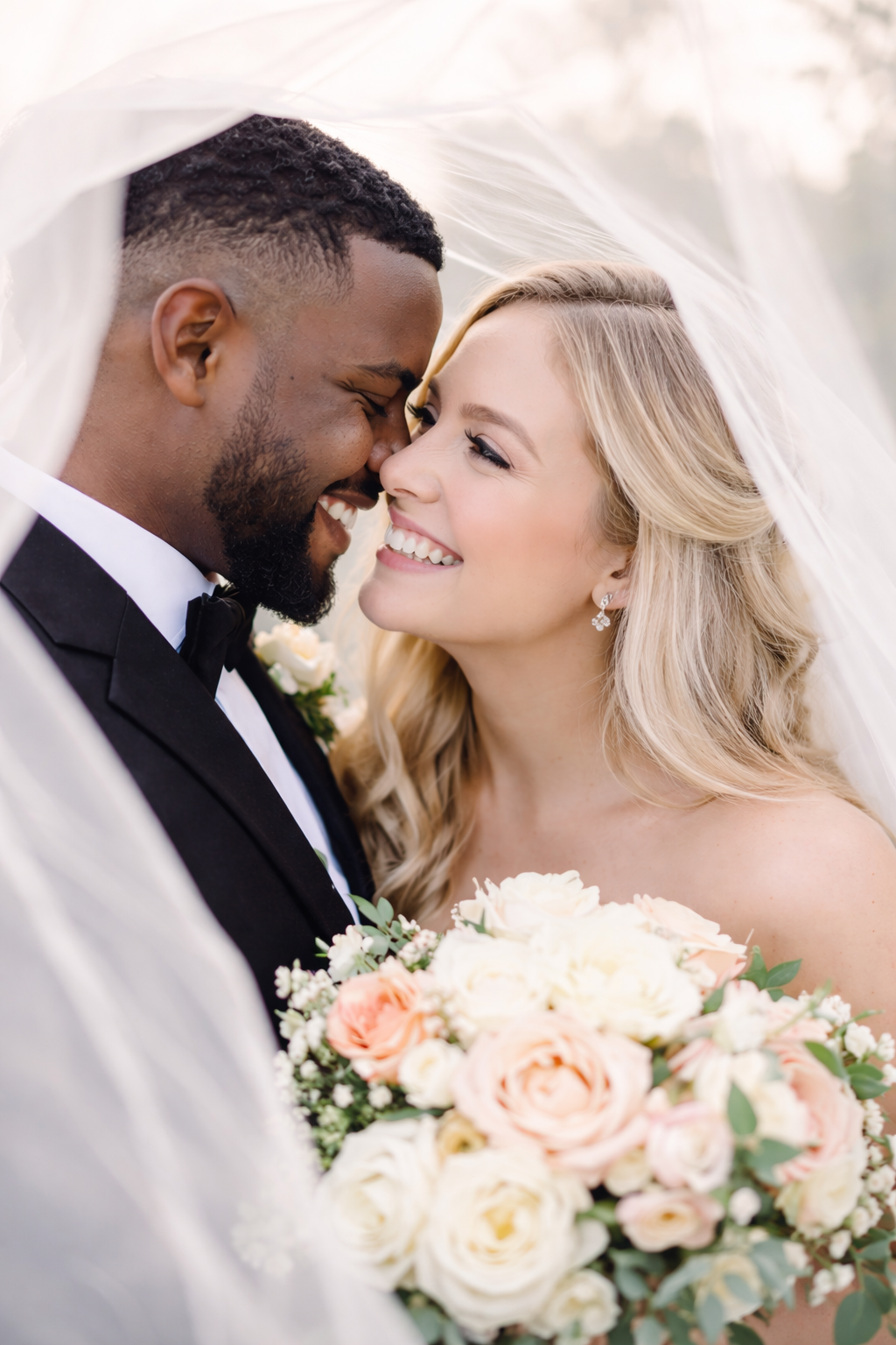 Happy newlywed couple shares an intimate moment under a veil with peach and white rose bouquet.