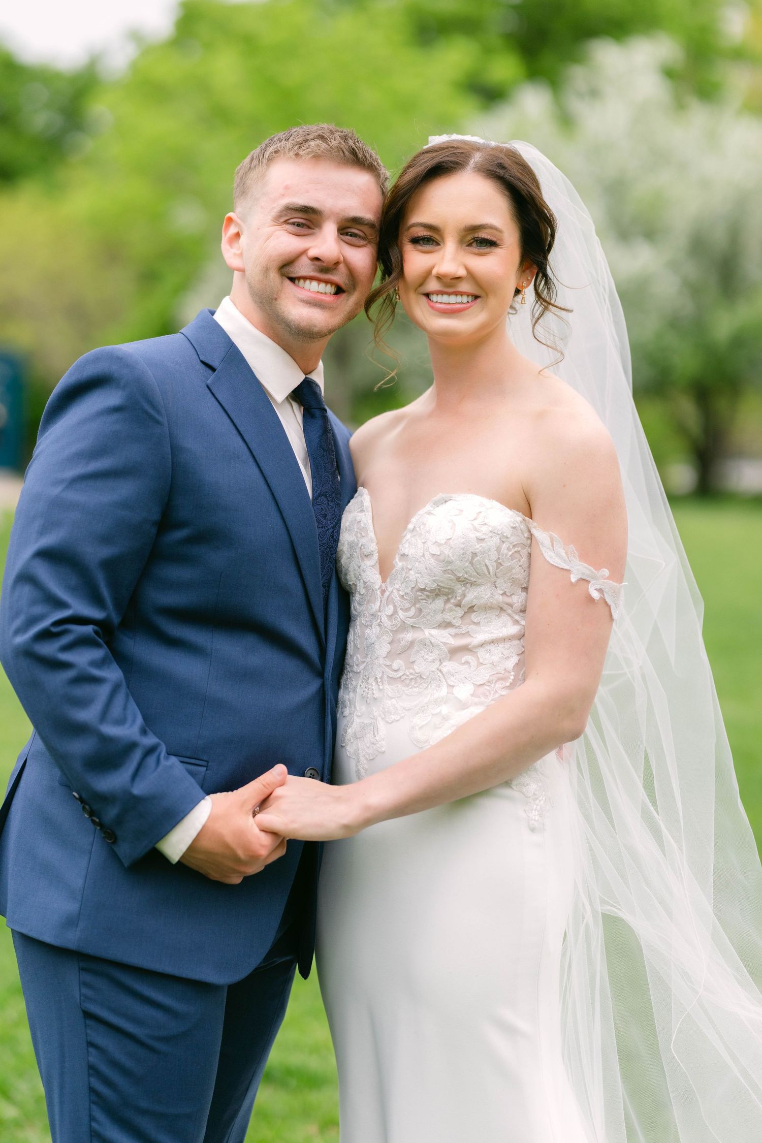 Smiling bride and groom hold hands in outdoor garden, bride wearing lace off-shoulder wedding gown.