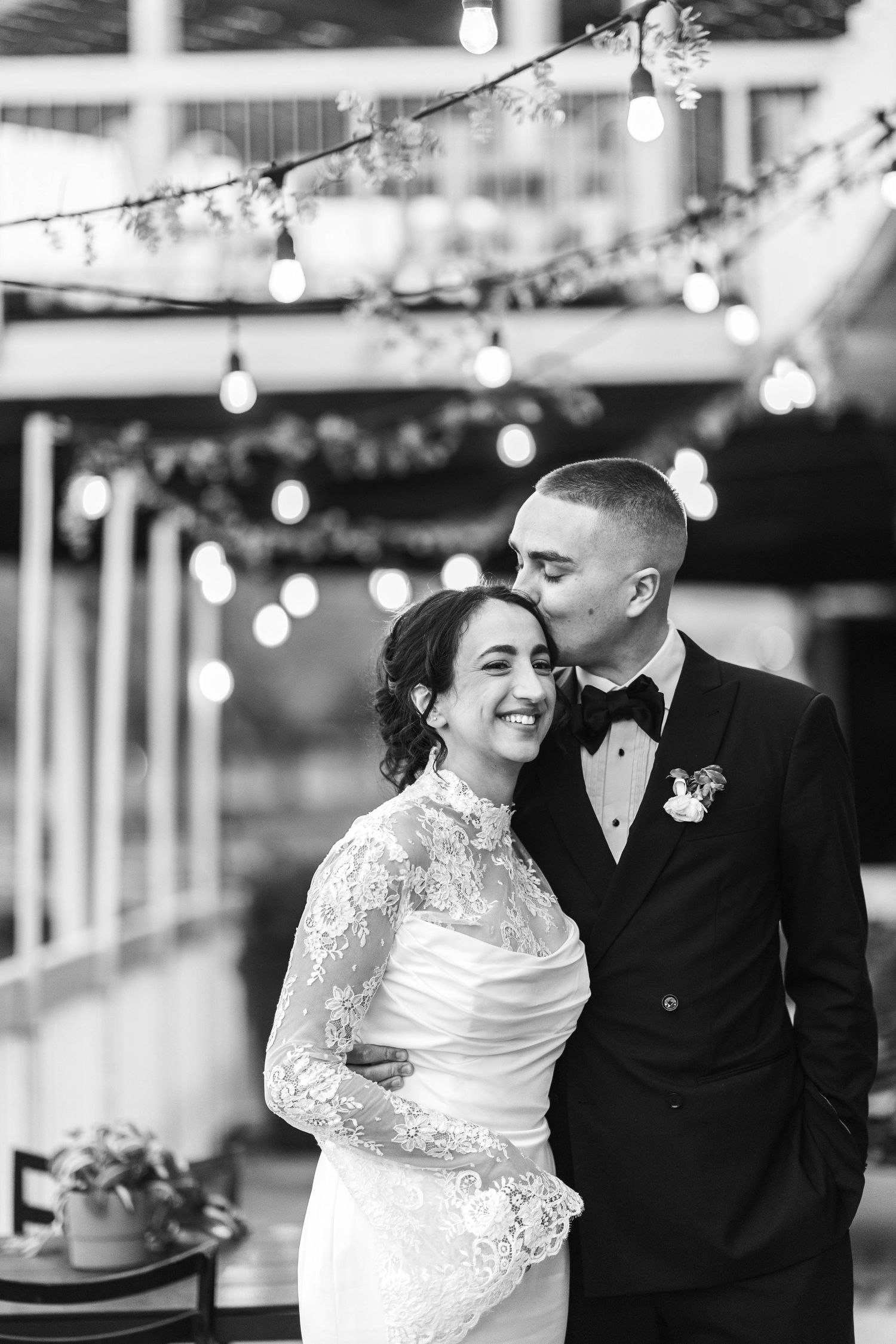 Black and white wedding portrait of smiling bride in lace gown and groom in tuxedo under string lights.