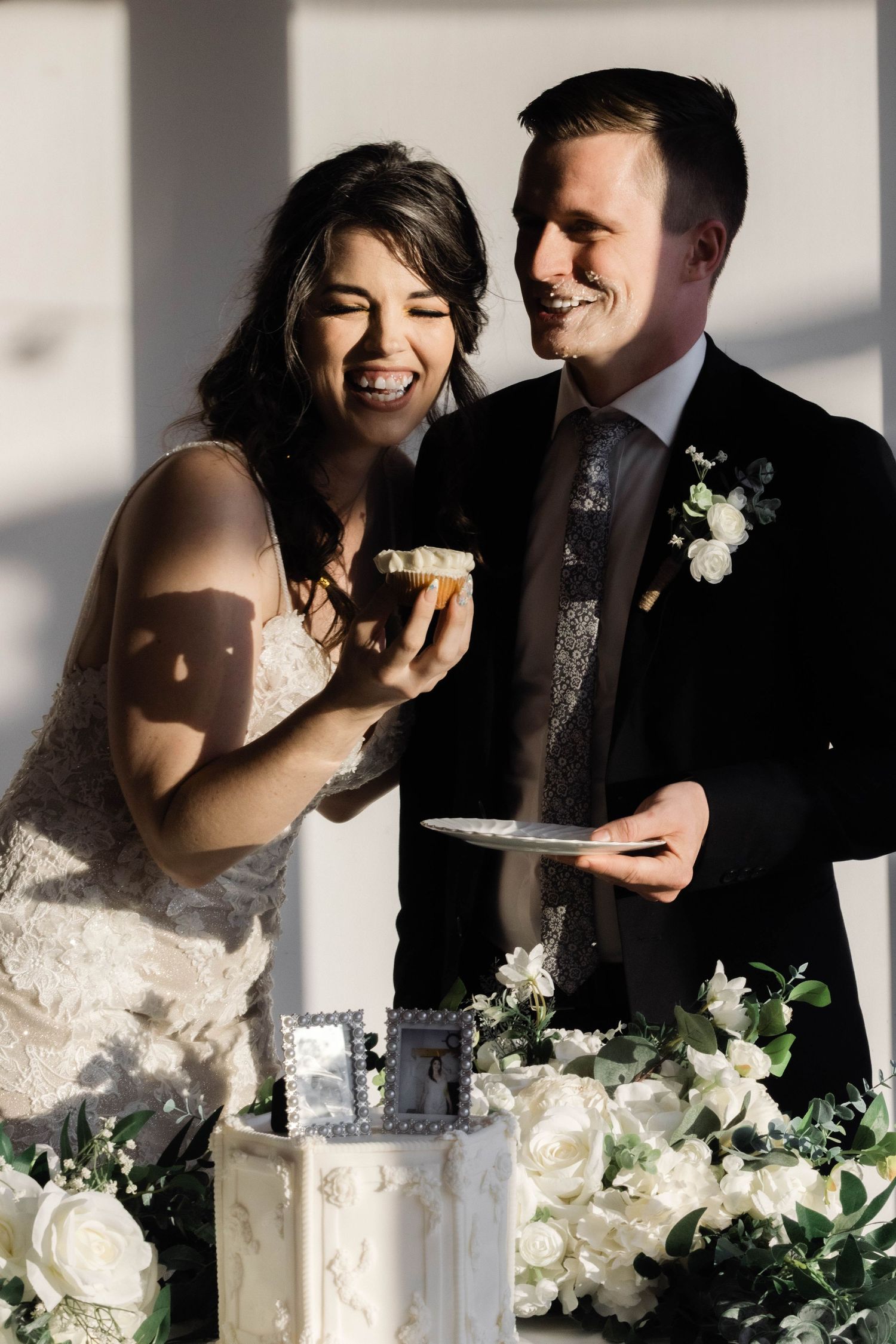 A joyful couple shares cake at their wedding reception surrounded by white floral decorations.
