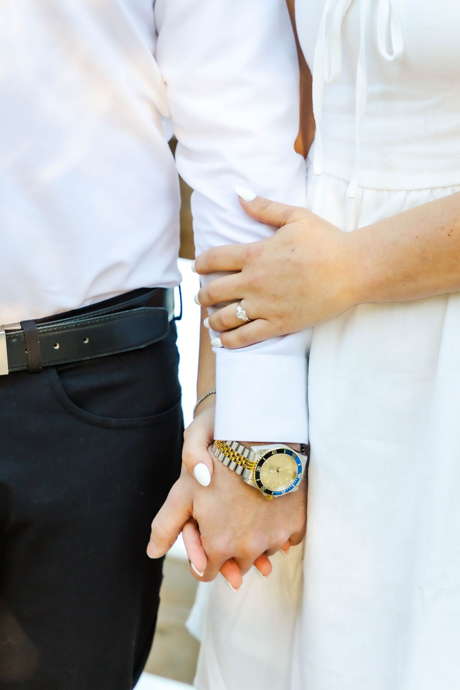 Close-up of couple holding hands with wedding rings and elegant watch visible against white and black formal attire.