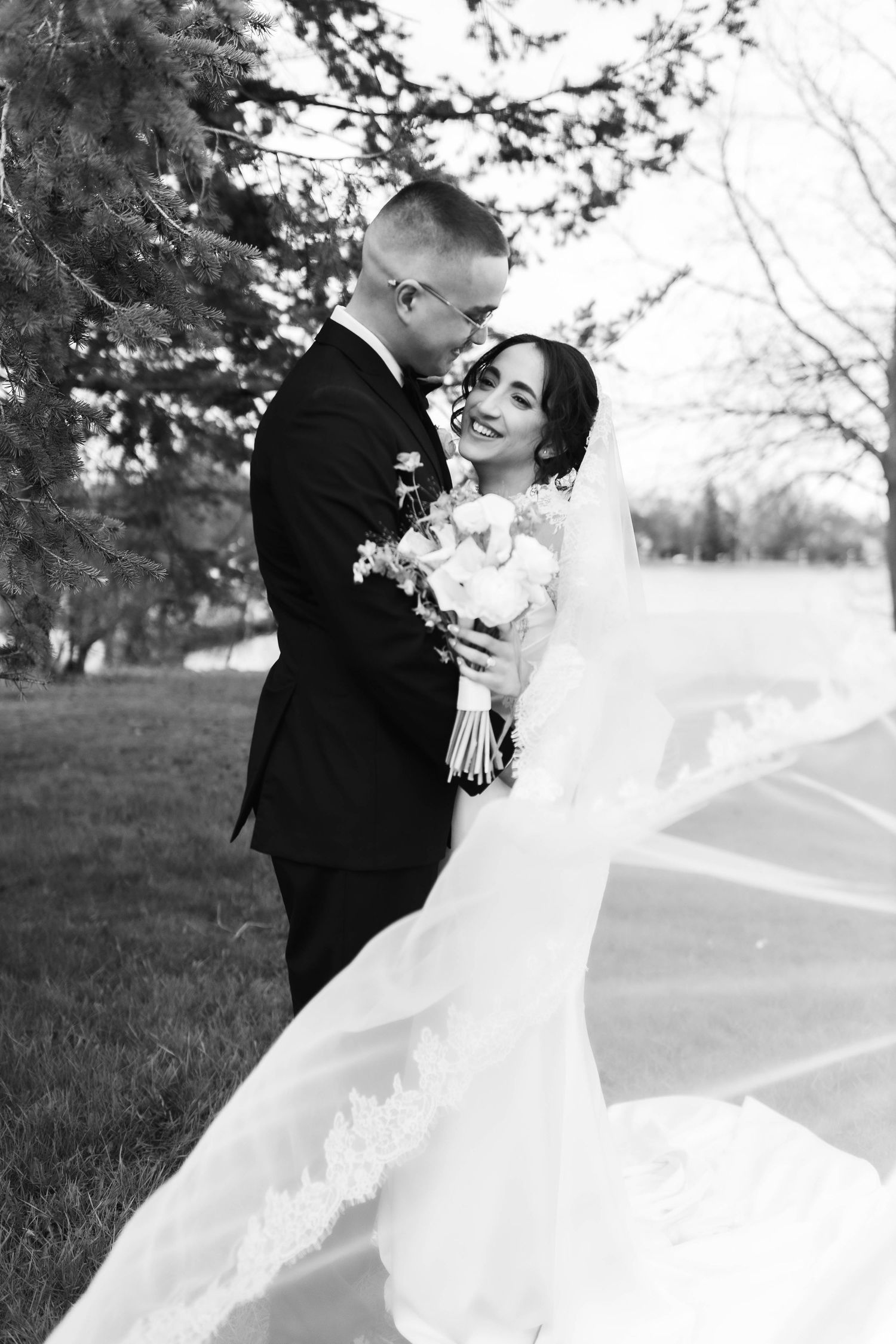 Black and white wedding photo of smiling bride in lace gown with flowing veil and groom in suit embracing outdoors.