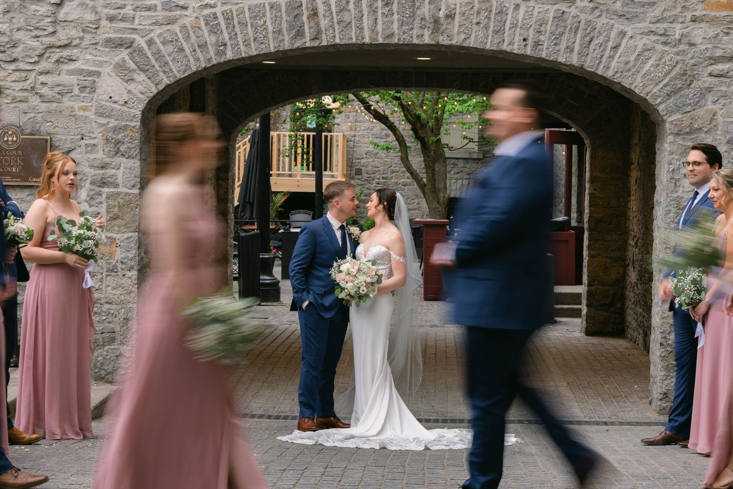 Wedding couple kissing under stone arch while bridal party in pink dresses walks past in motion blur.