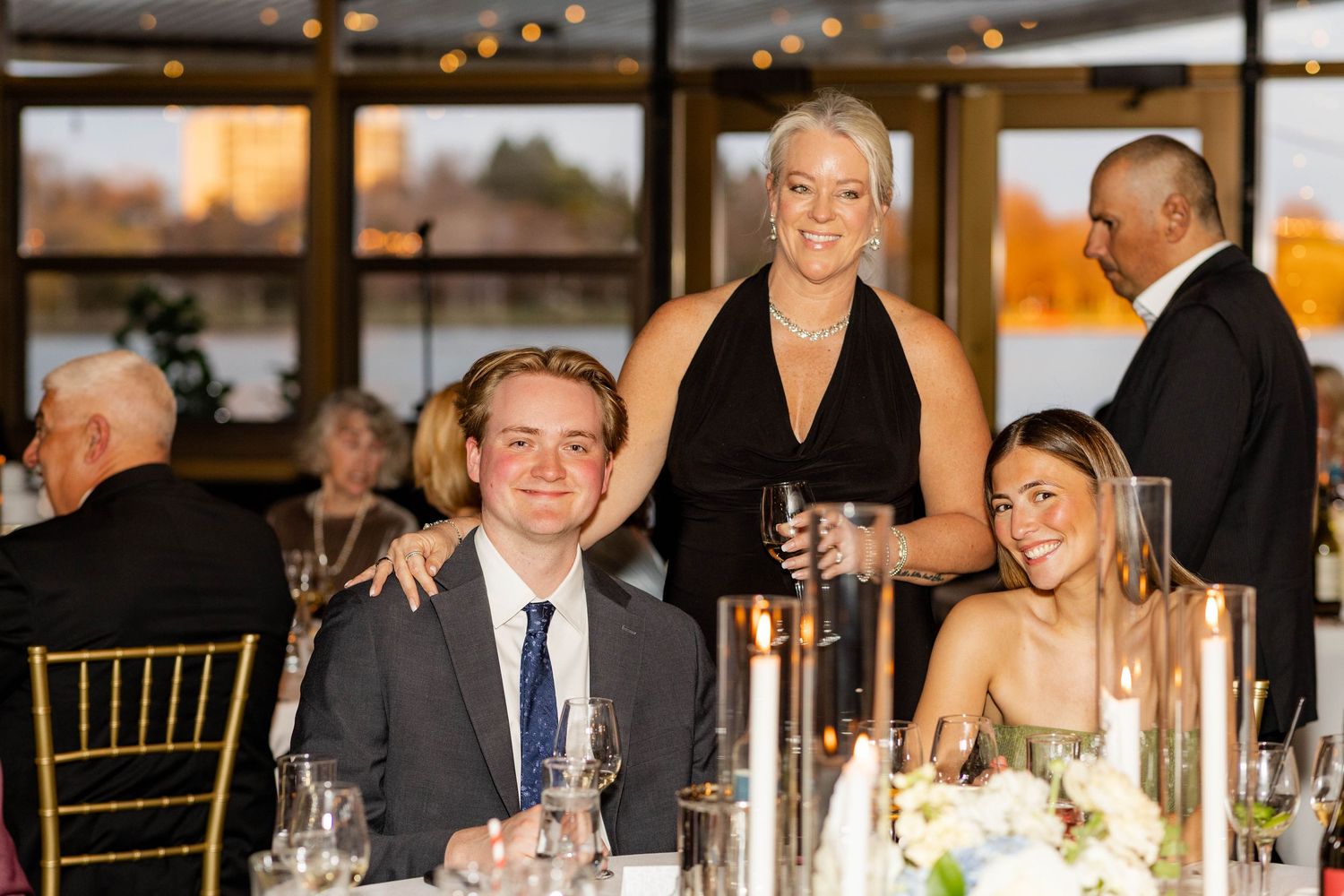 Three elegantly dressed guests smiling at a wedding reception dinner table in a waterfront venue.