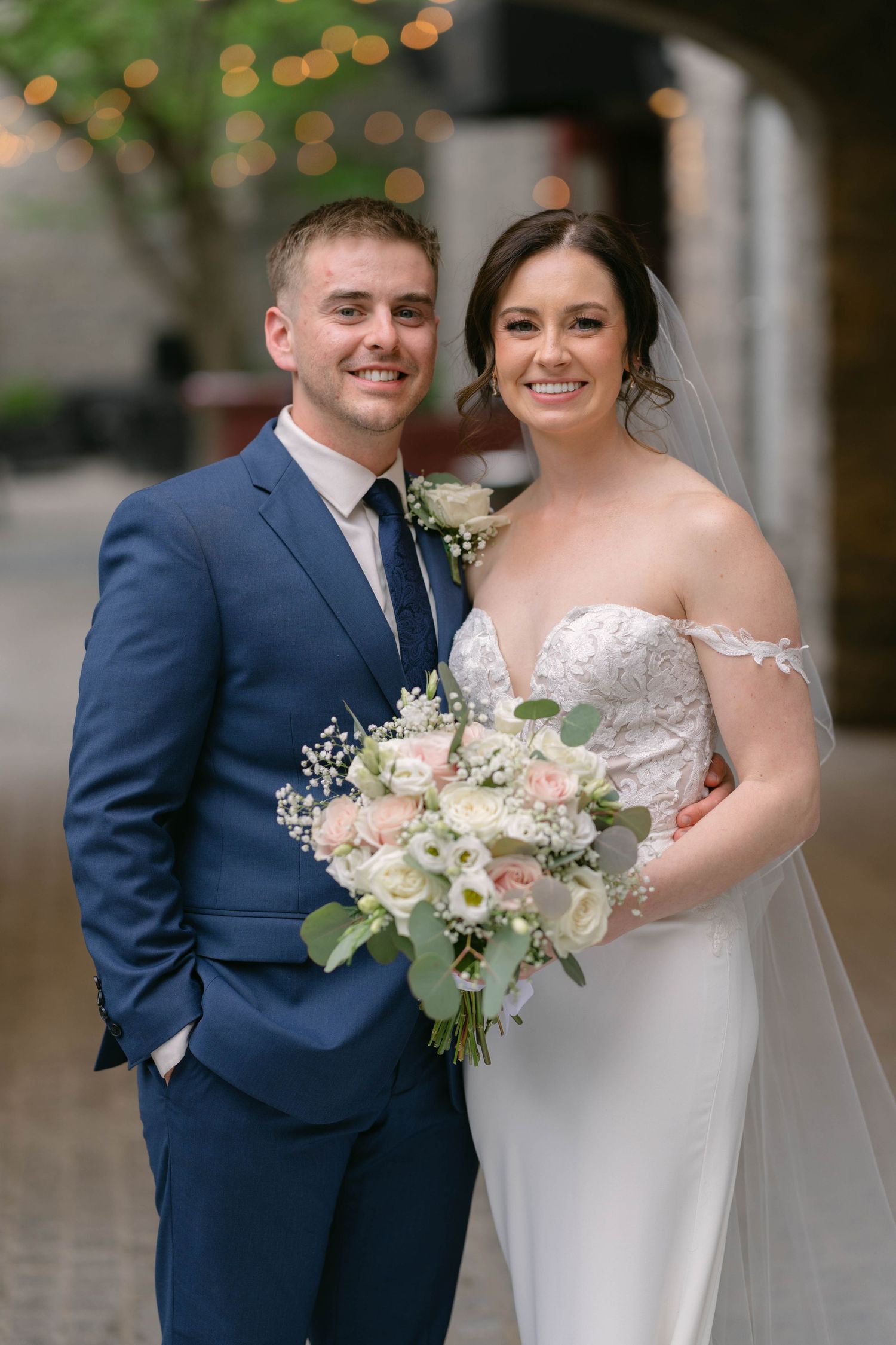 Smiling bride and groom pose together outdoors, she holds a pink and white bouquet, he wears a navy suit.