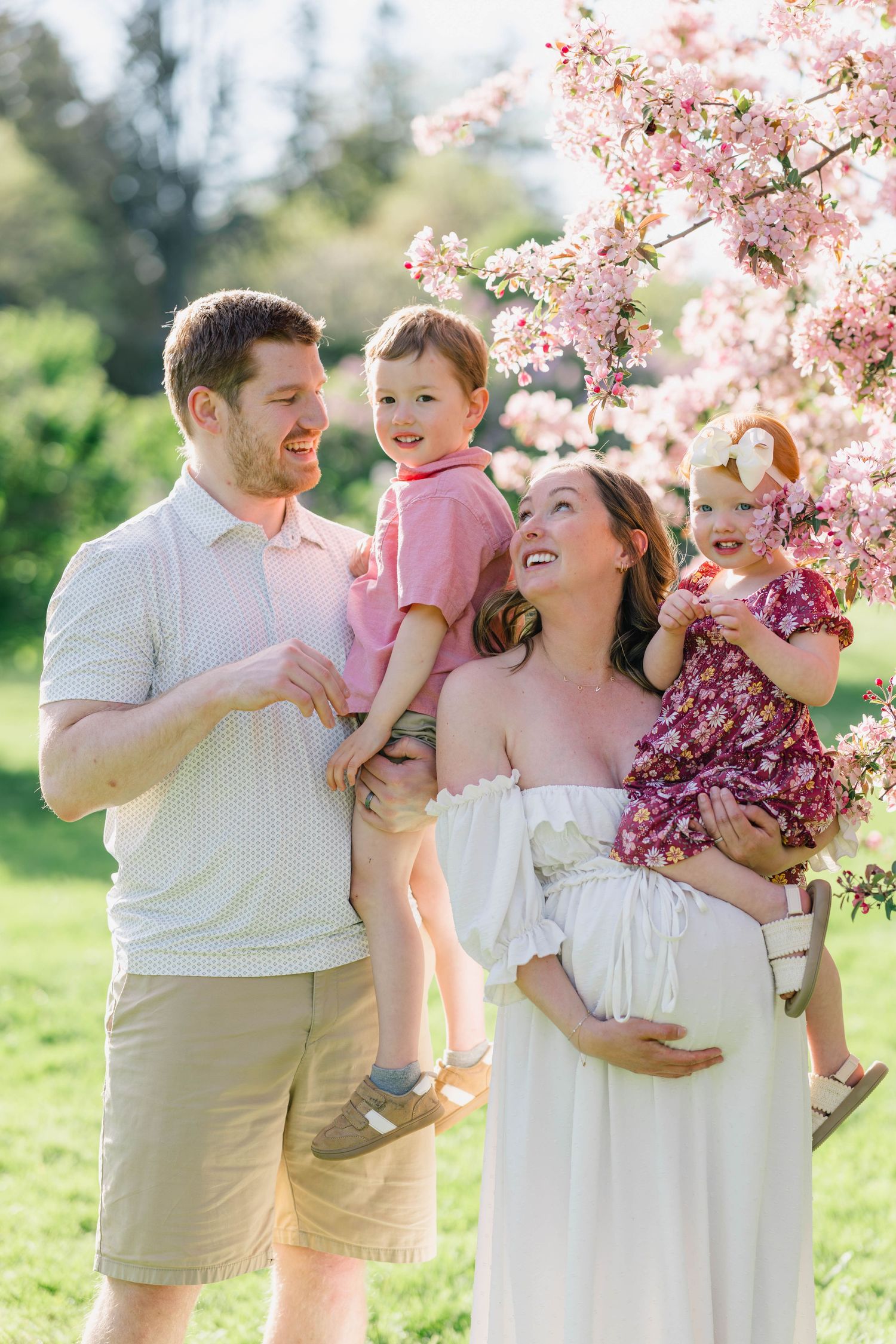 Pregnant woman in white dress with husband and two young children under pink cherry blossom tree outdoors.
