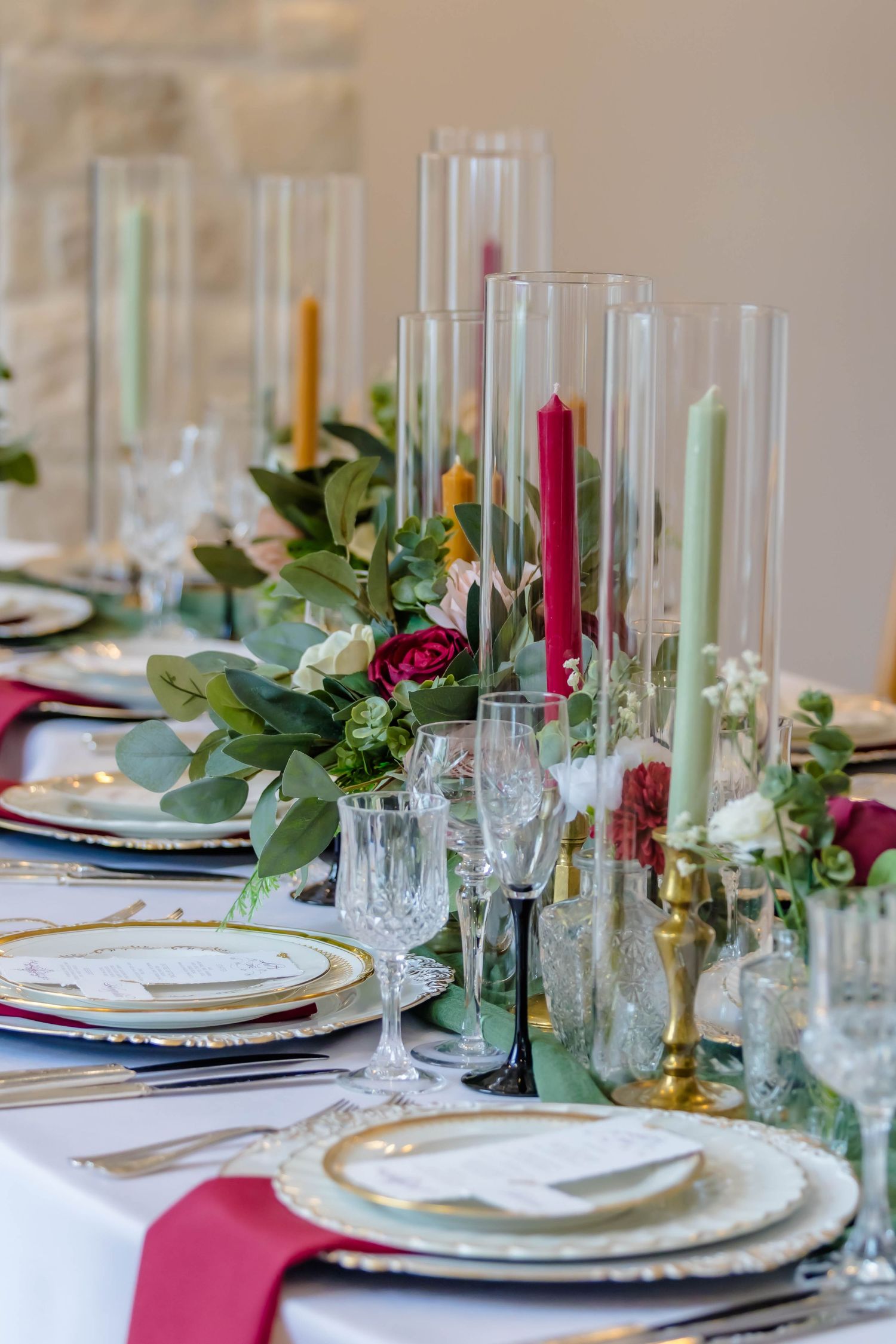 Elegant table setting with tall glass candle holders, red taper candles, white plates, and eucalyptus greenery centerpiece.