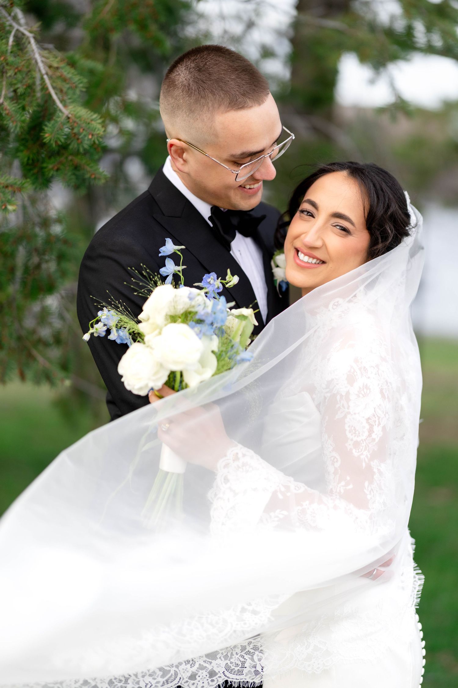 Bride and groom embrace outdoors, her lace dress and veil flowing, holding white and blue bouquet under pine trees.