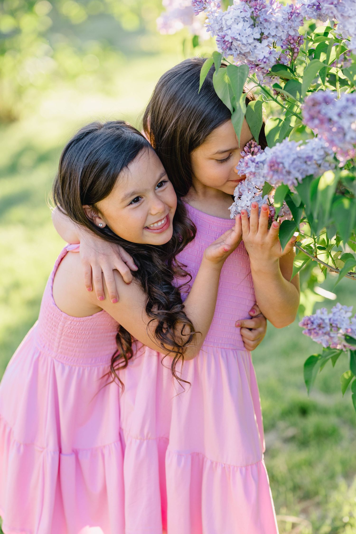 Two young girls in matching pink dresses smile and smell lilac flowers outdoors in a sunny garden.