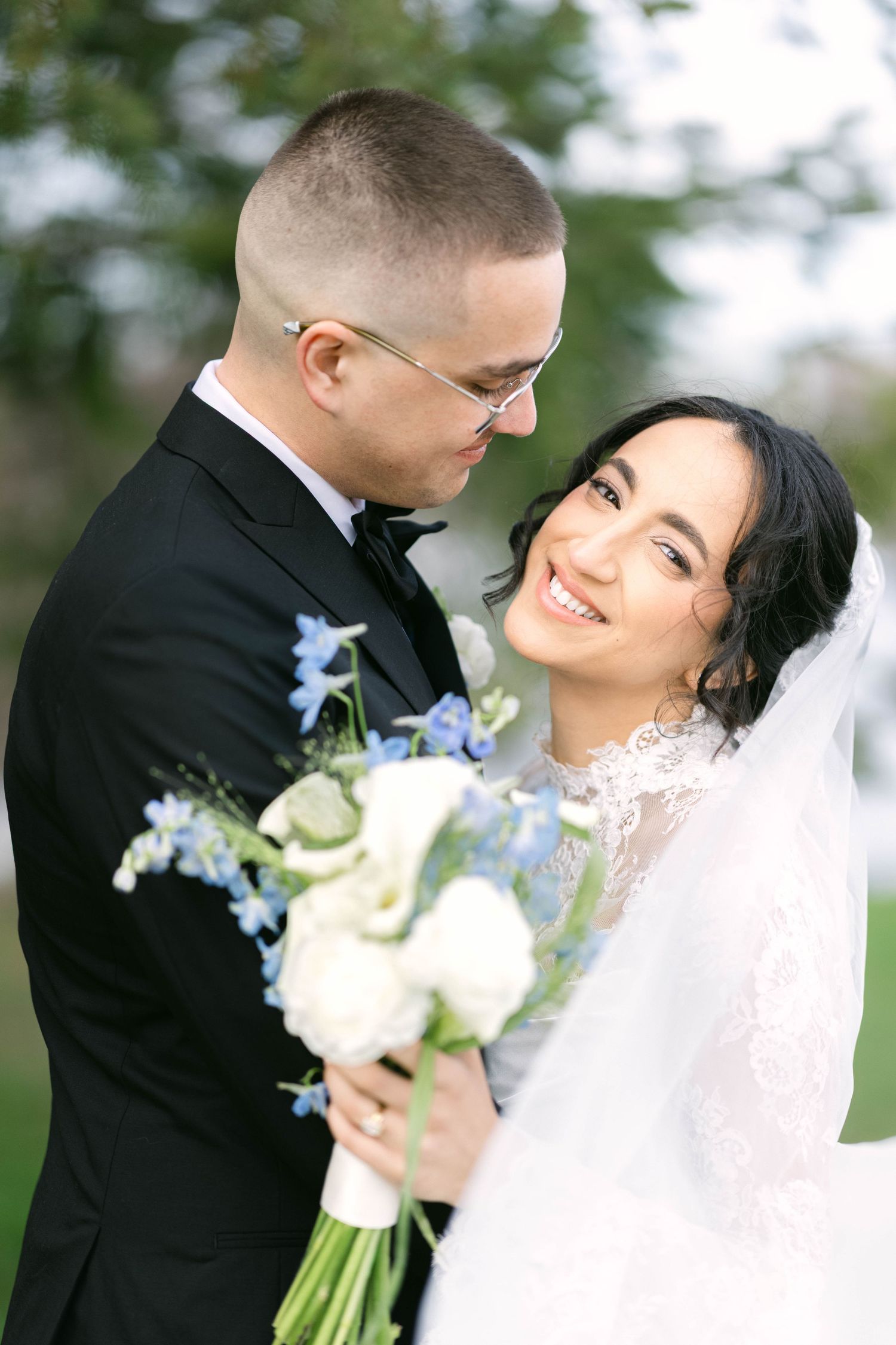 Smiling bride in lace gown holds white and blue bouquet as groom in black tuxedo leans in close outdoors.
