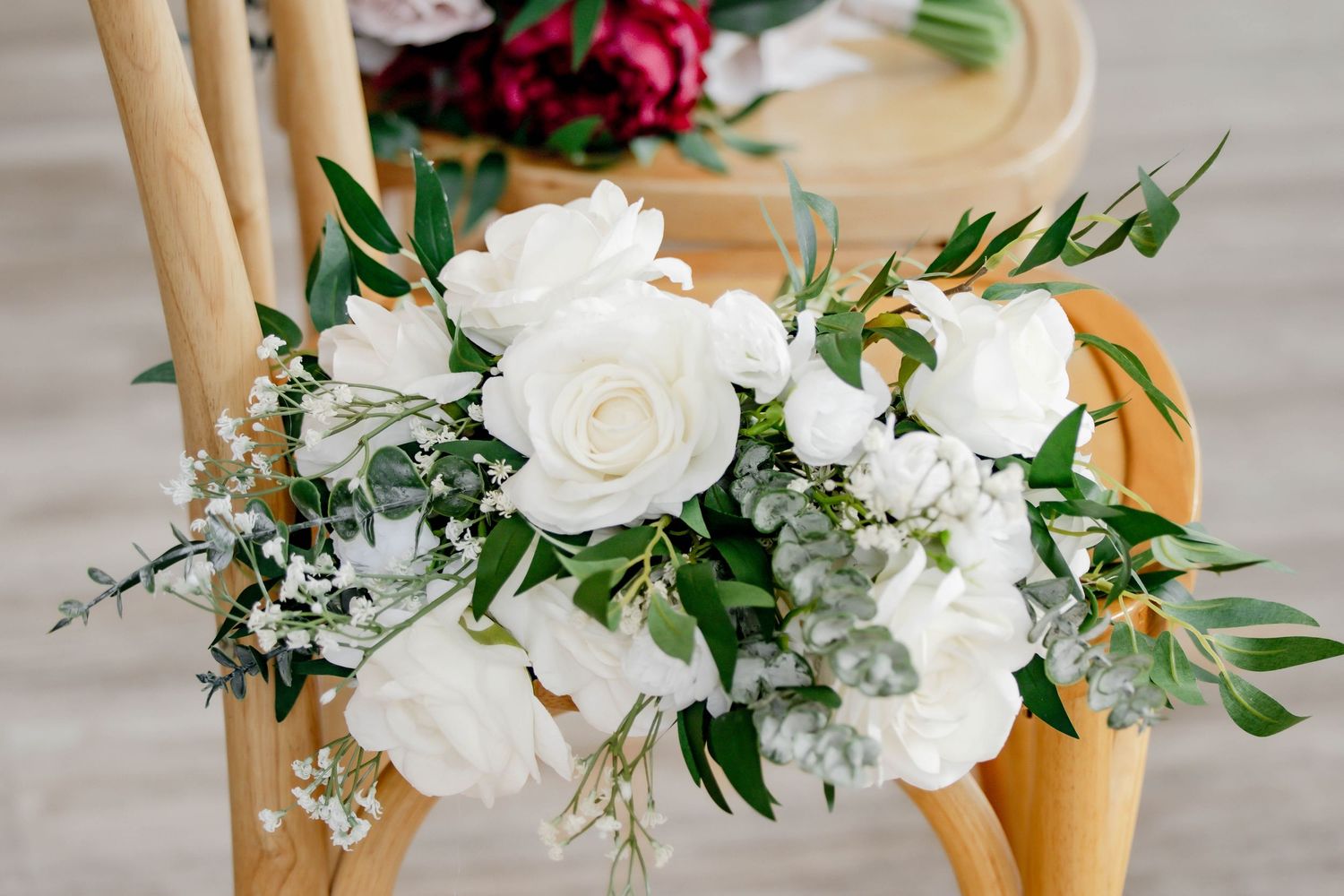 Delicate white floral arrangement with roses and greenery adorns wooden chairs at a wedding ceremony.