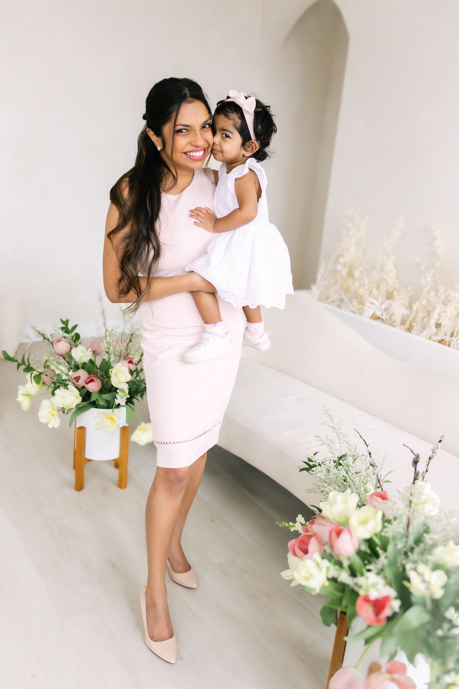 Mother and child in matching pink outfits share tender moments against white backdrop with floral decorations.