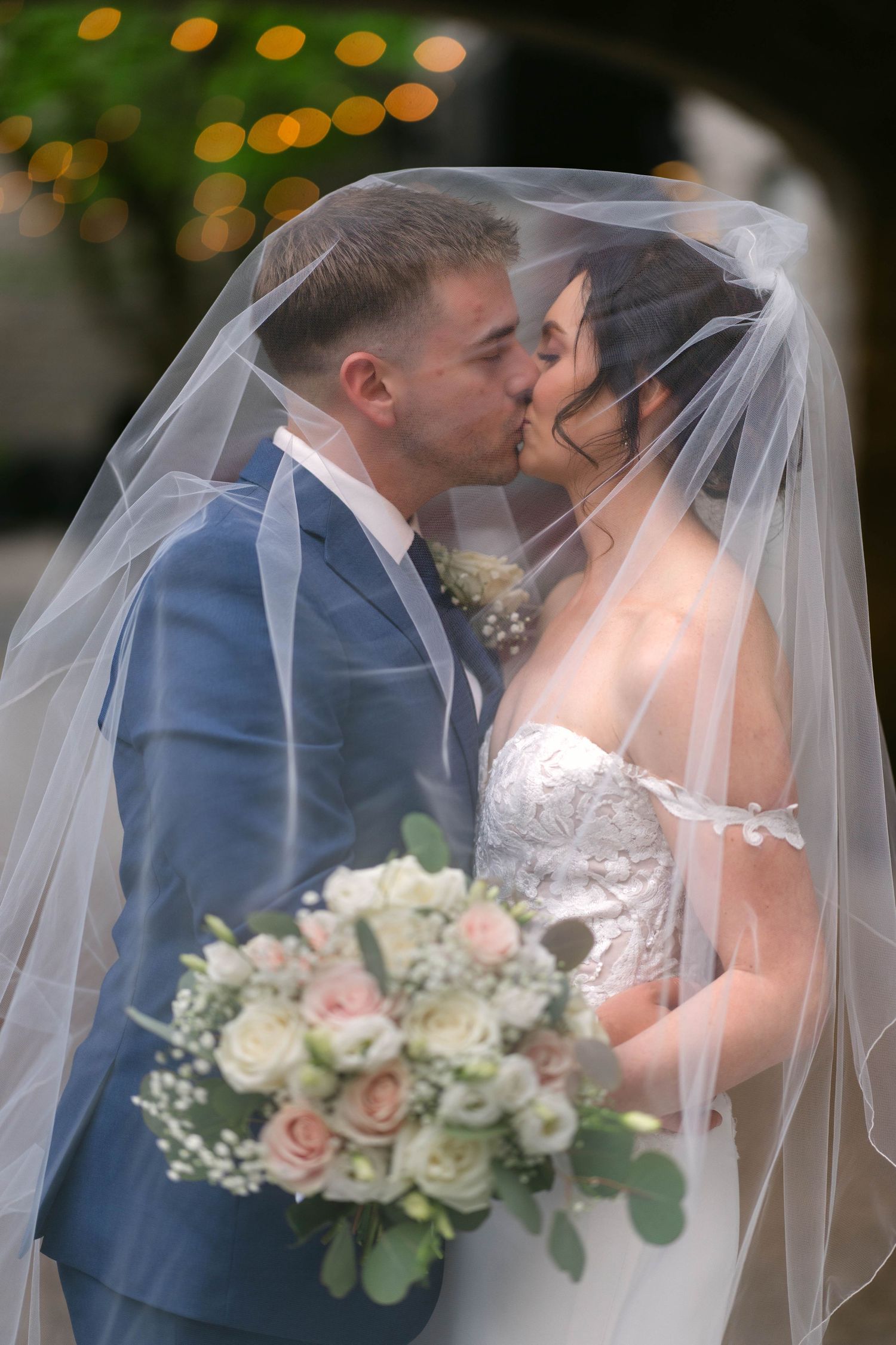 Bride and groom sharing a kiss under a white veil, bride holding a pink and white rose bouquet.