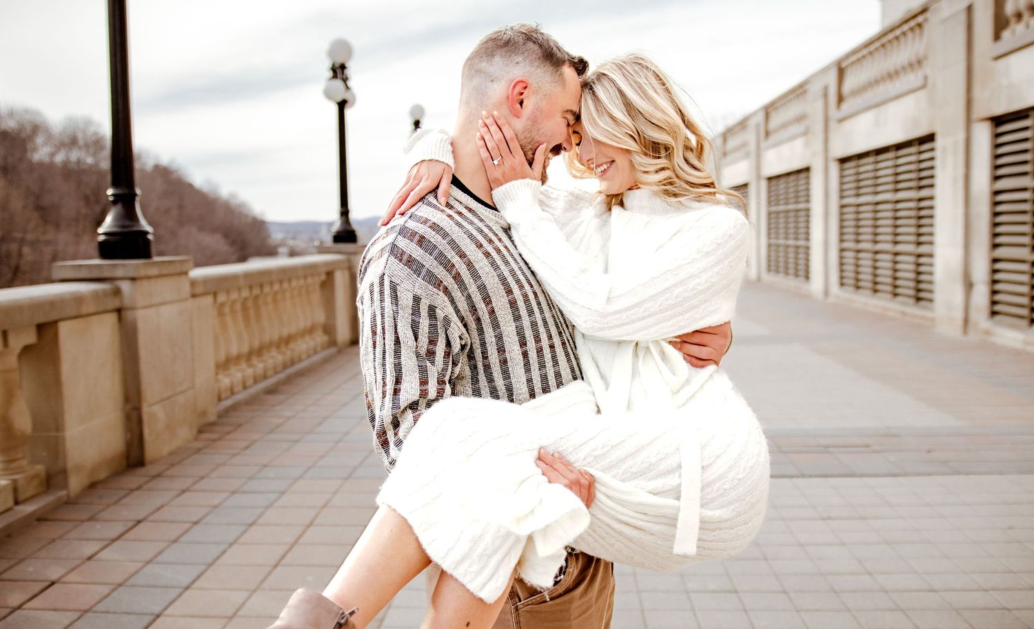 A romantic moment captured on a city bridge as a couple shares an intimate embrace in warm sunlight.