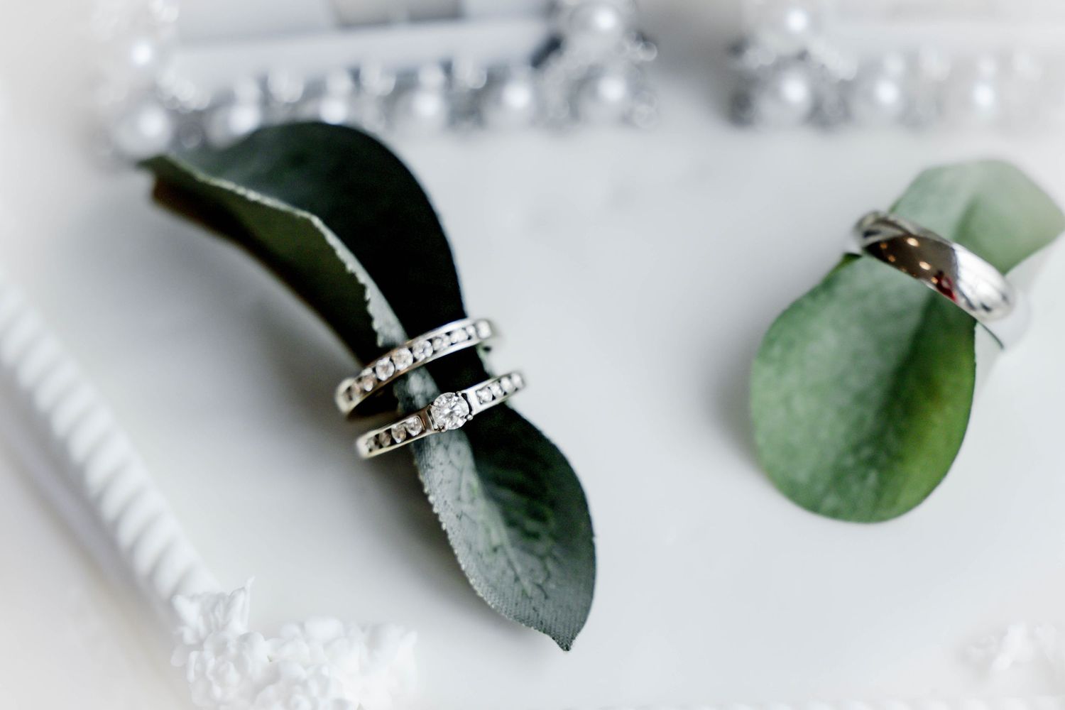 Wedding rings rest elegantly on green eucalyptus leaves against a white background.