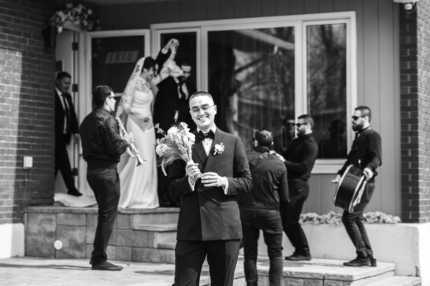 Groom in black tuxedo holds bouquet outside church as wedding party gathers on steps in black and white photo.
